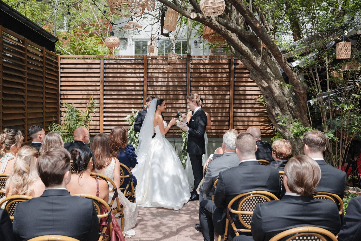 Outdoor wedding ceremony with bride and groom exchanging vows before seated guests, surrounded by lush trees and a wooden backdrop.