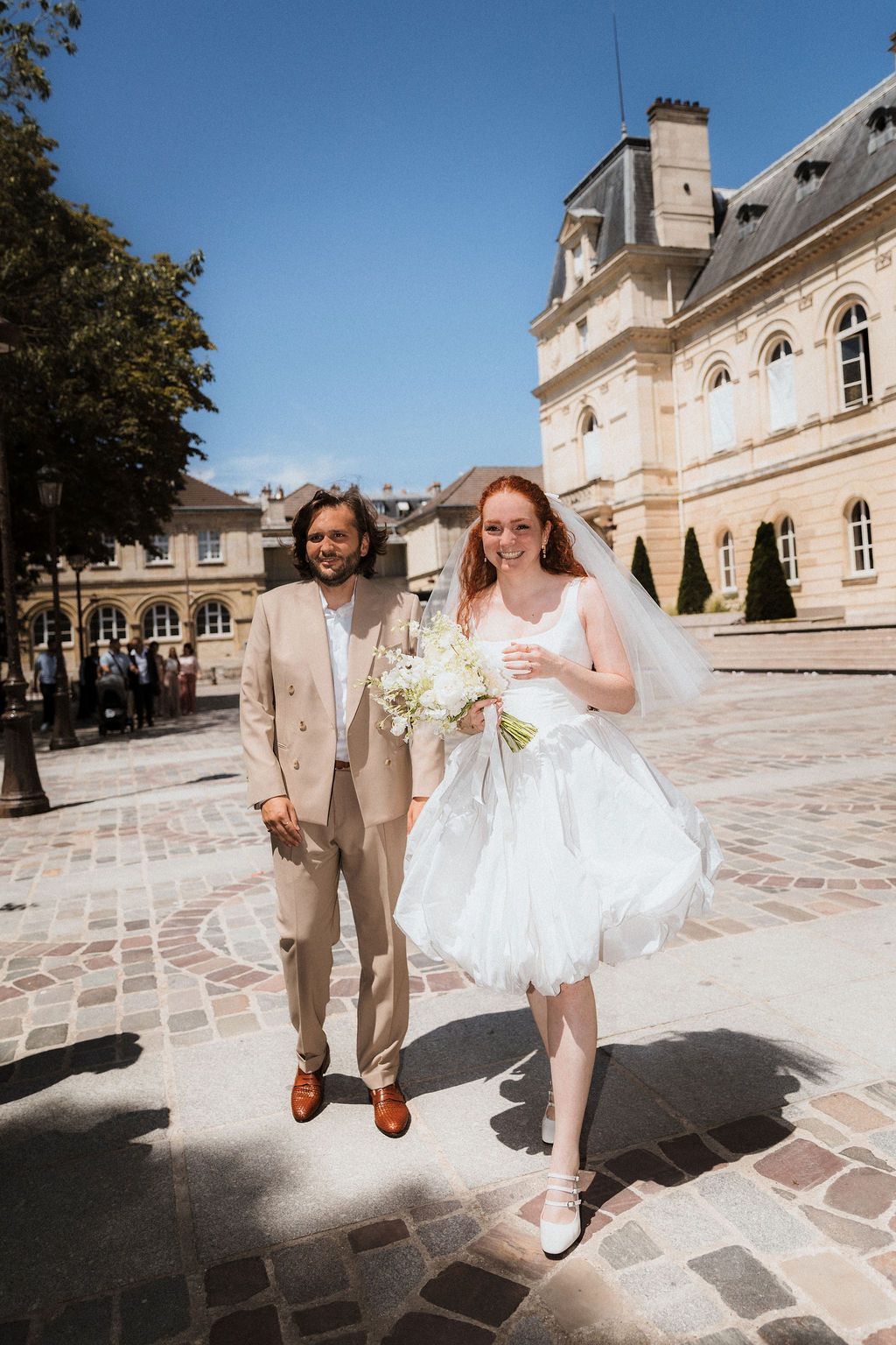 Newlyweds pose playfully on stone courtyard of elegant French chateau during wedding photo session.