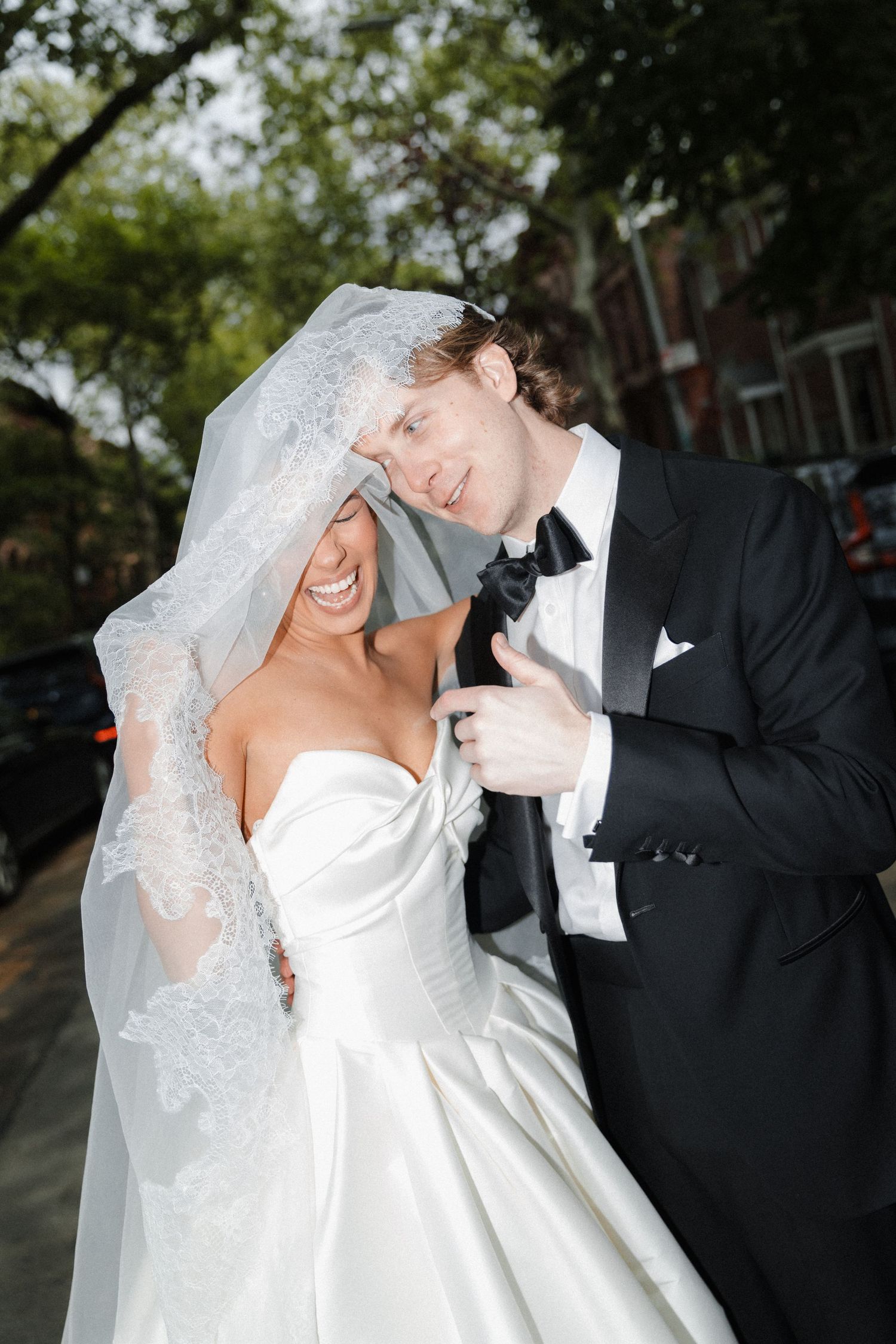 Joyful newlyweds share a laugh outdoors, bride in strapless gown with lace veil, groom in classic black tuxedo.