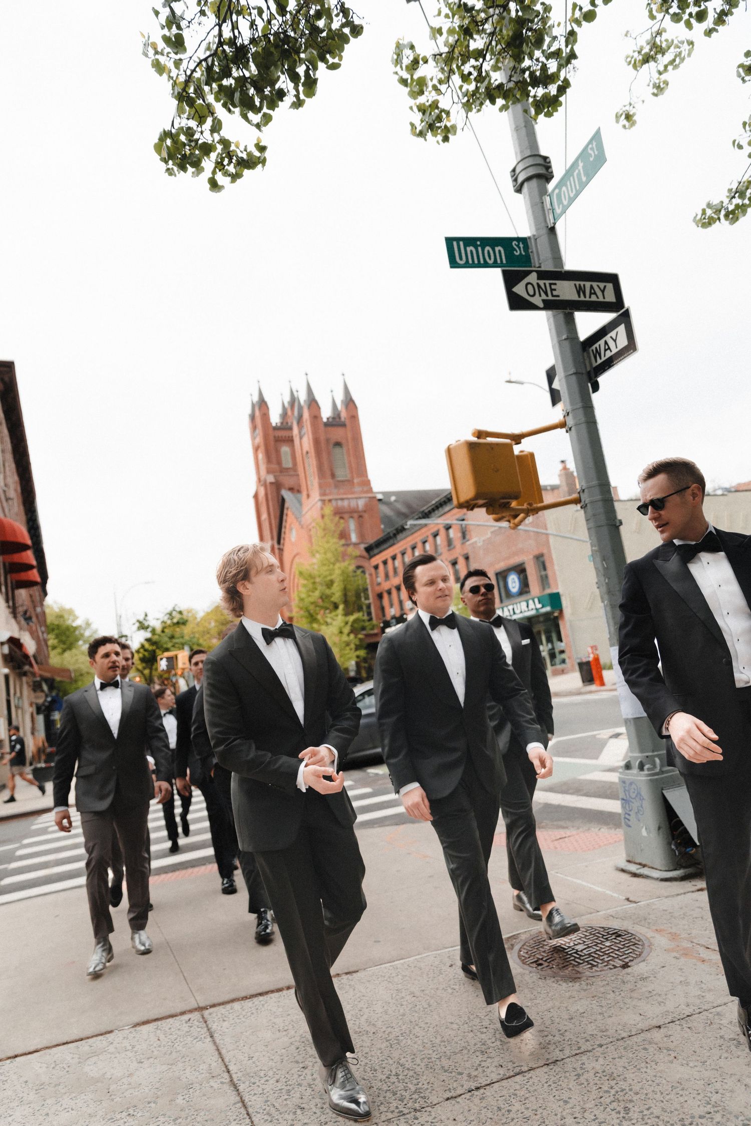Groomsmen in black tuxedos walking on Union St in Brooklyn, NYC, with a historic church in the background.