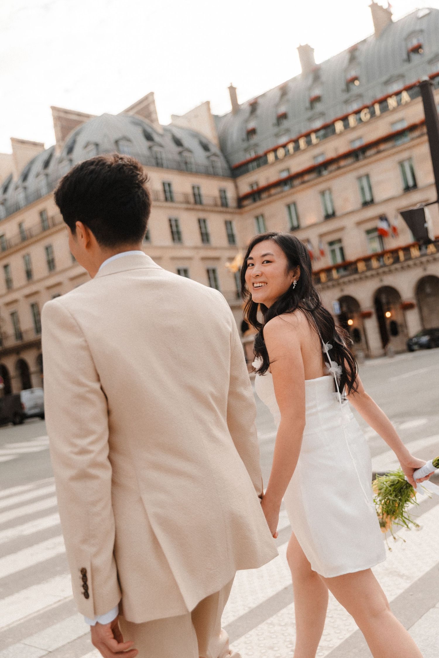 Asian couple in wedding attire holding hands crossing street in Paris near Hotel Regina.
