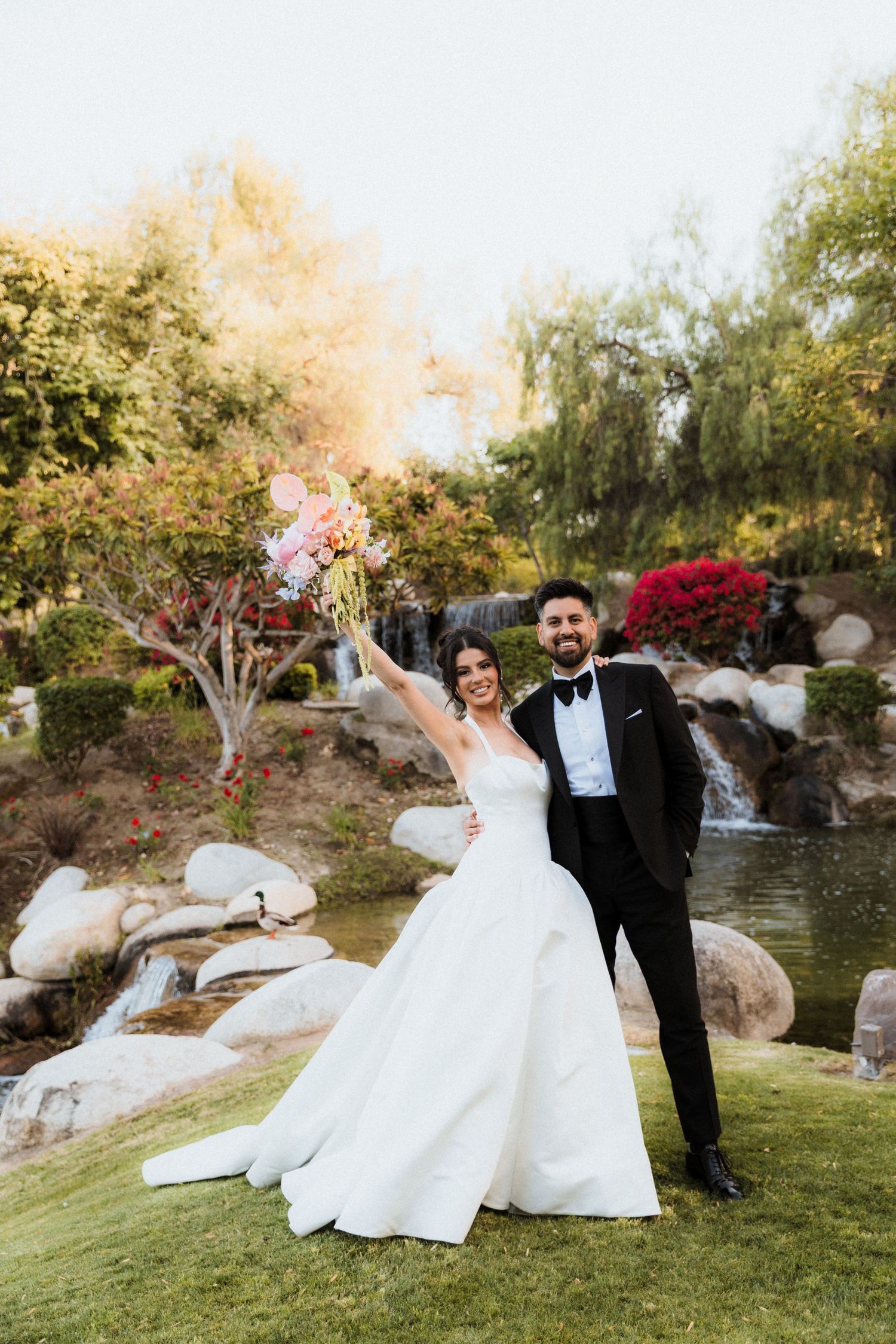 Bride and groom celebrate outdoors in garden with waterfall, she raises pink bouquet in white gown, he wears black tuxedo.