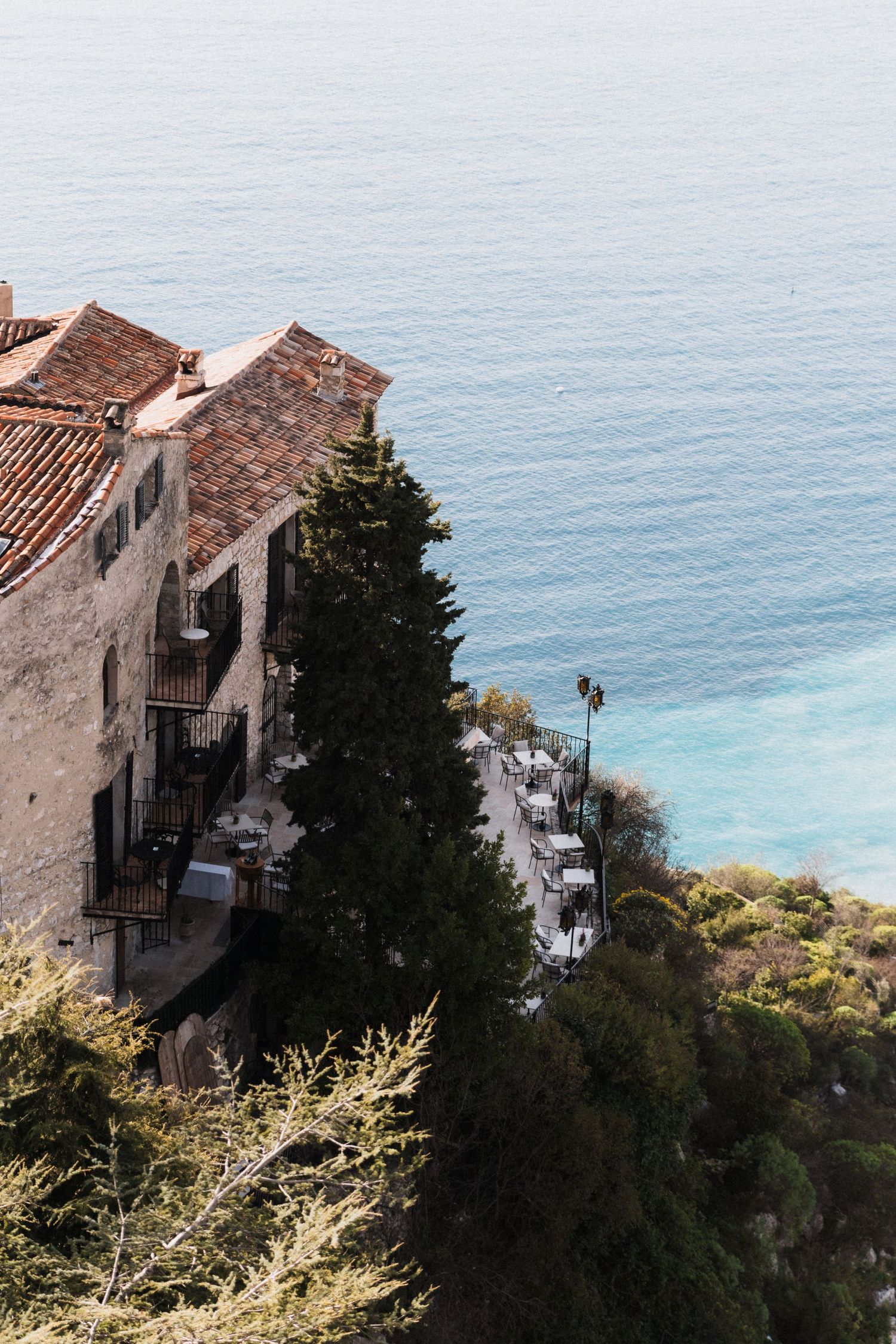 Cliffside Mediterranean Château Eza with terracotta roof overlooking turquoise sea, surrounded by lush green trees.