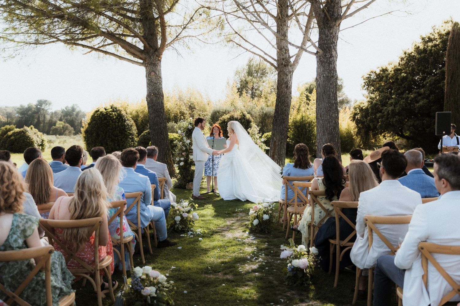 Outdoor wedding ceremony setup with white chairs and guests under tall trees.