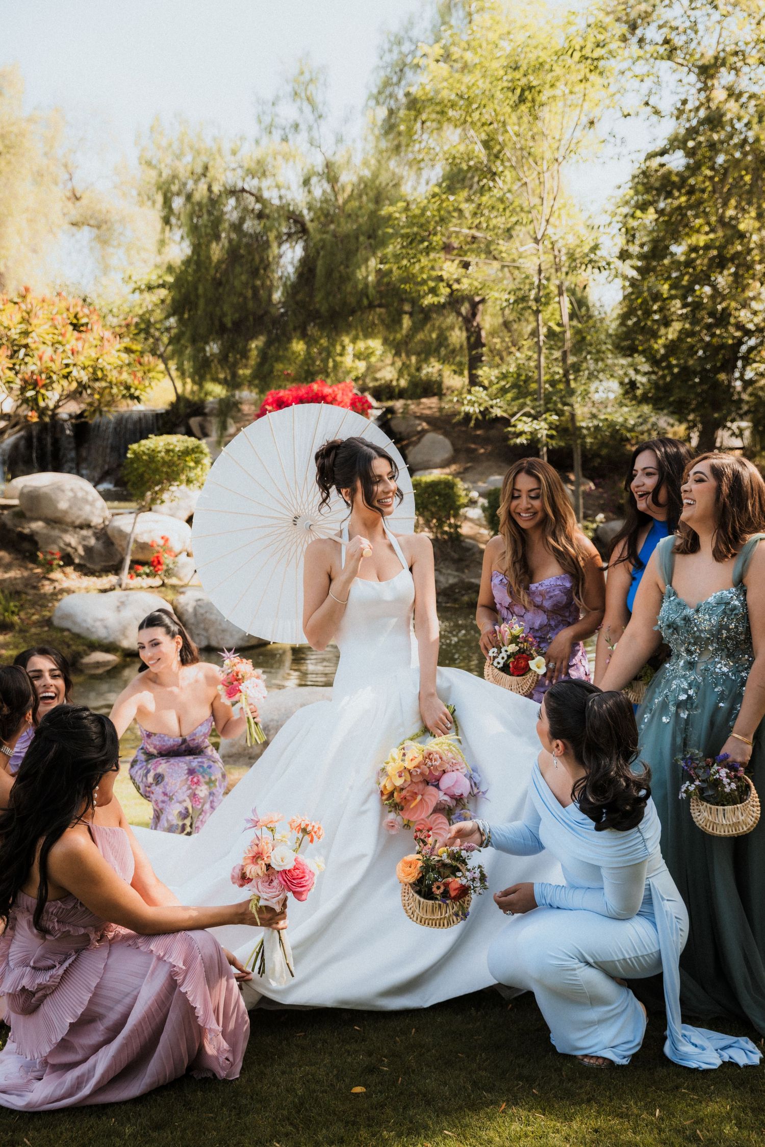 Bride holding white parasol surrounded by bridesmaids in colorful dresses in a garden setting.