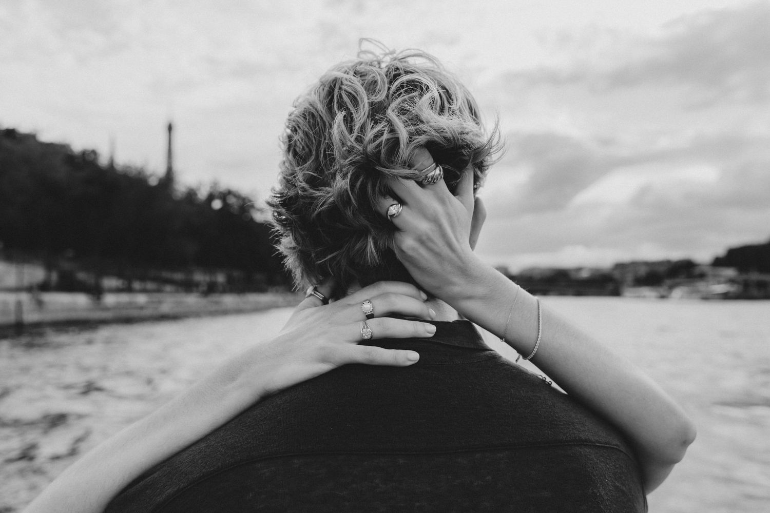 Black and white photo of a woman with curly hair touching her neck, wearing a bracelet, with the Eiffel Tower in background.
