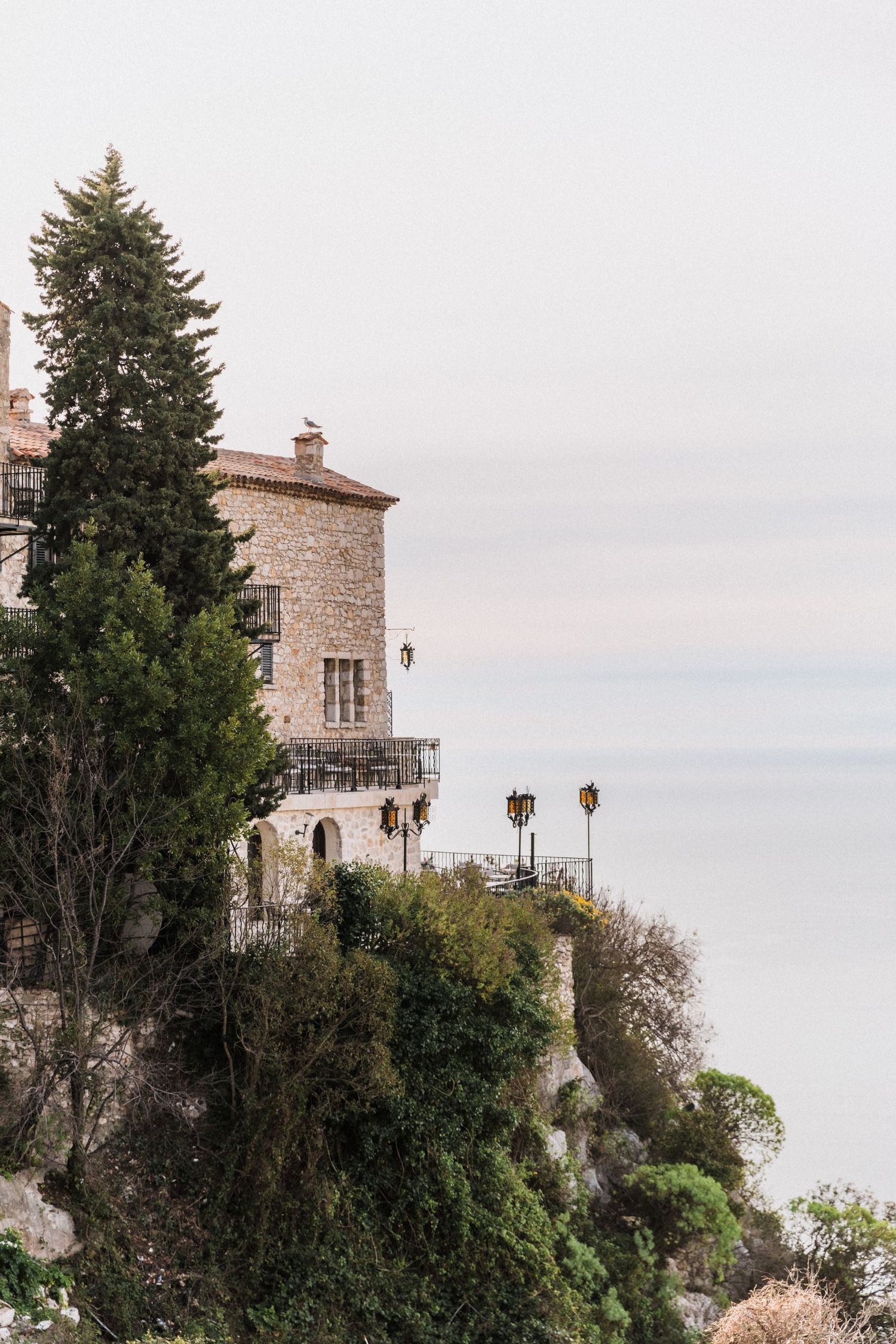 Château Eza - Historic stone castle perched on a cliff overlooking a misty sea, surrounded by tall cypress trees and lush greenery.