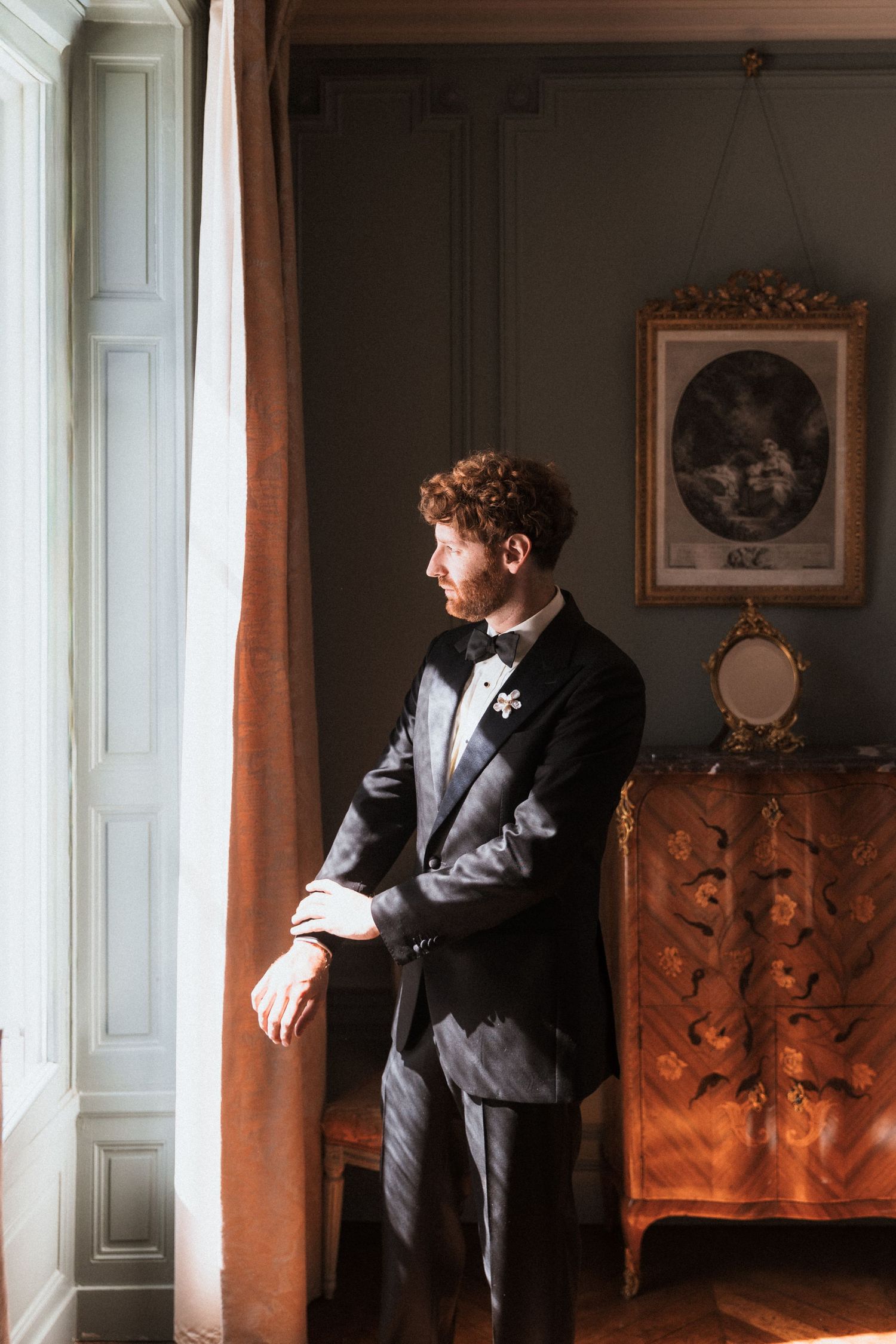 Elegant groom in black tuxedo and bow tie stands by window in ornate vintage room with antique furniture.