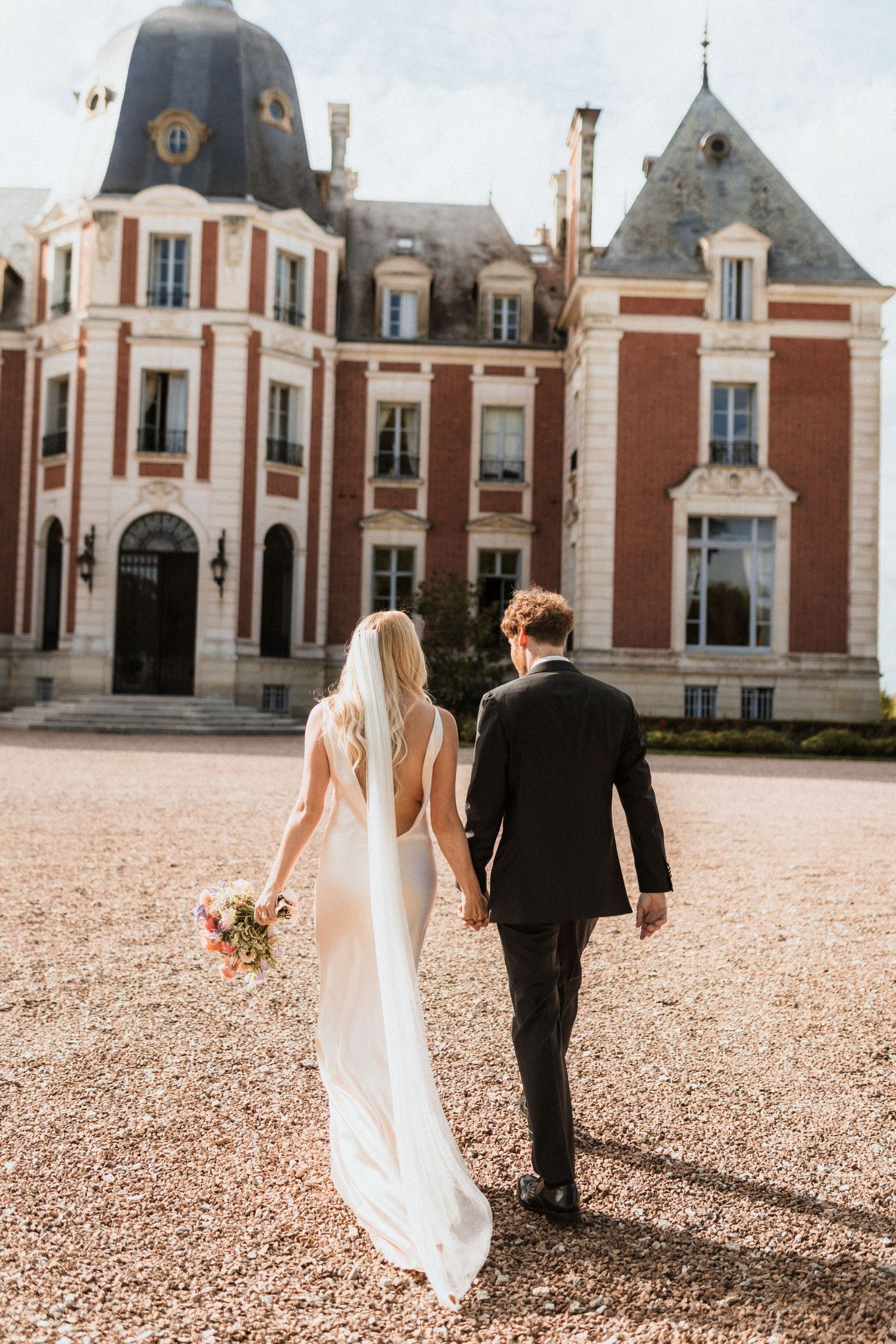 Bride and groom walking hand in hand toward a grand French château on their wedding day.
