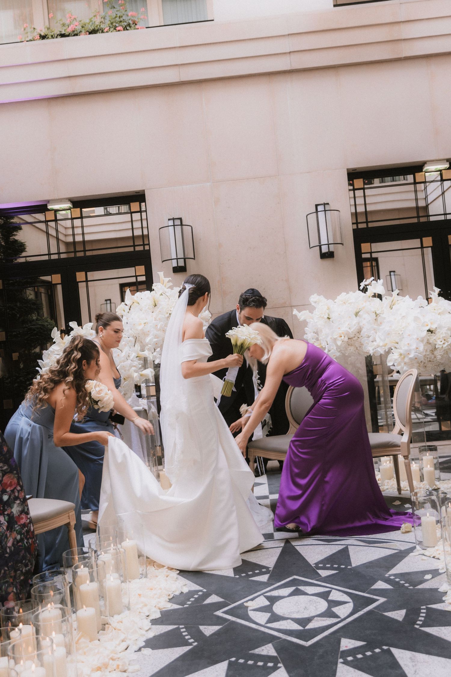 Wedding party members help arrange a flowing white bridal train on a decorative black and white patterned floor.