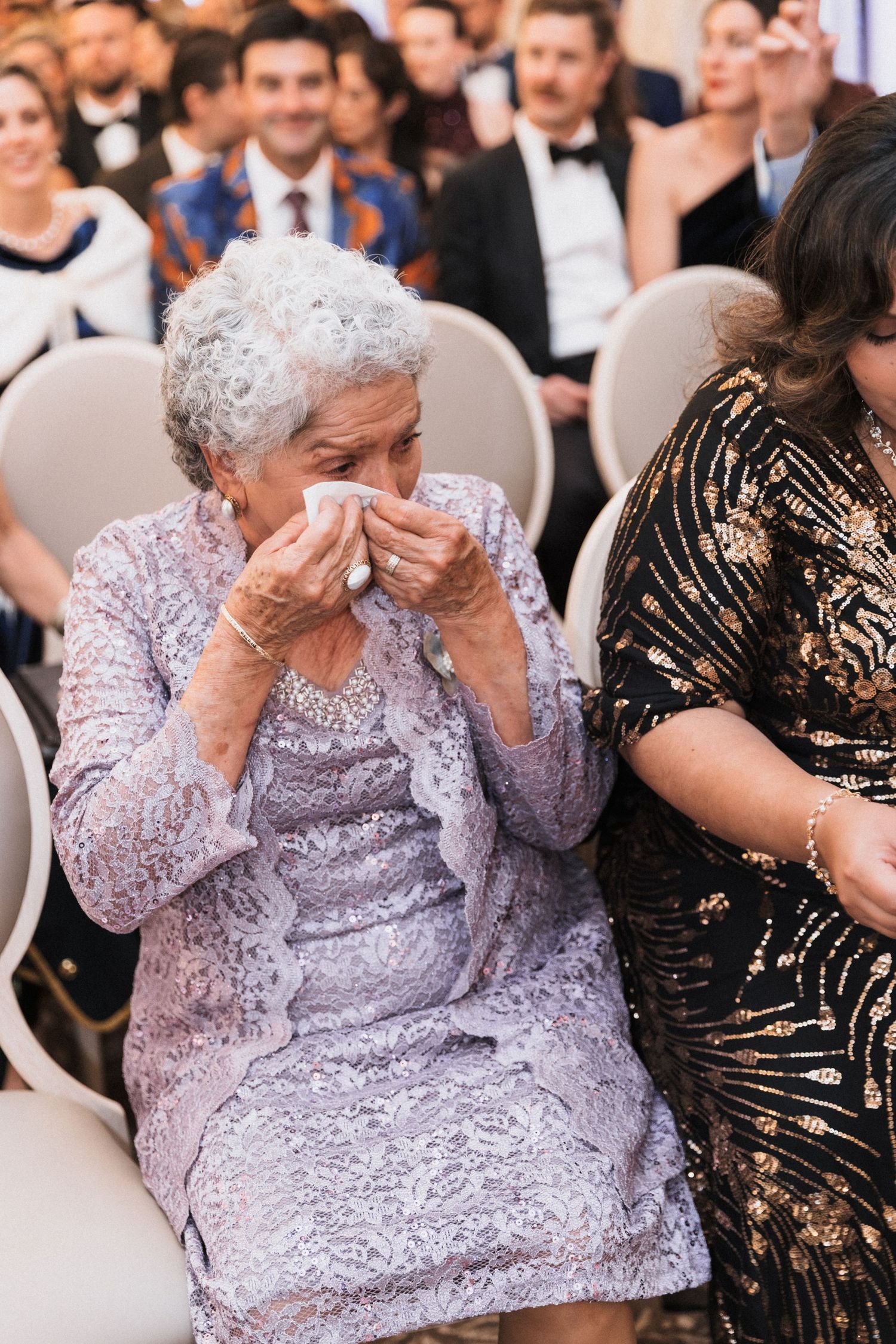 Elderly woman in lavender lace dress dabbing tears with tissue at a wedding ceremony.