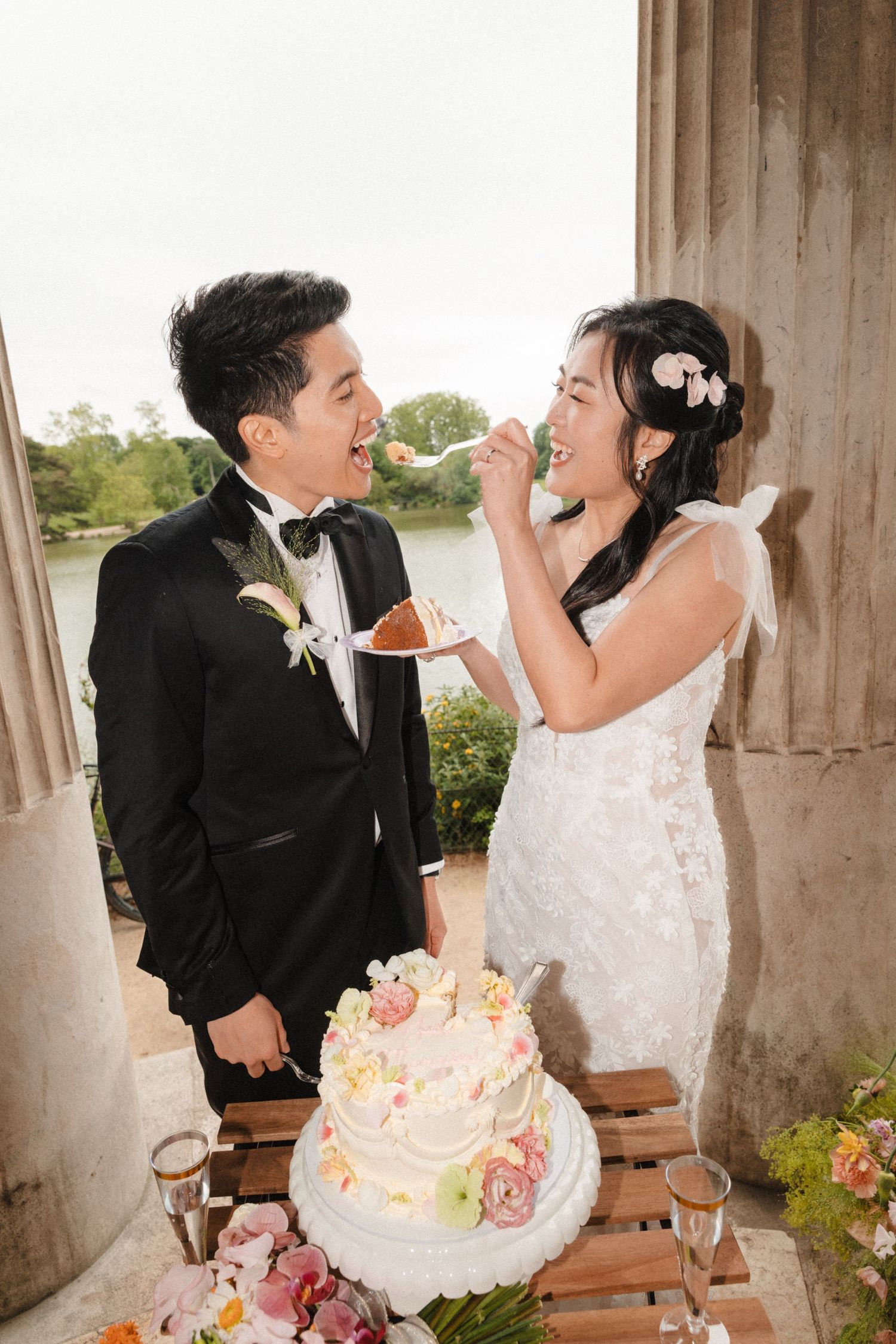 A bride and groom feed each other wedding cake outdoors near a lake, smiling joyfully in elegant wedding attire.