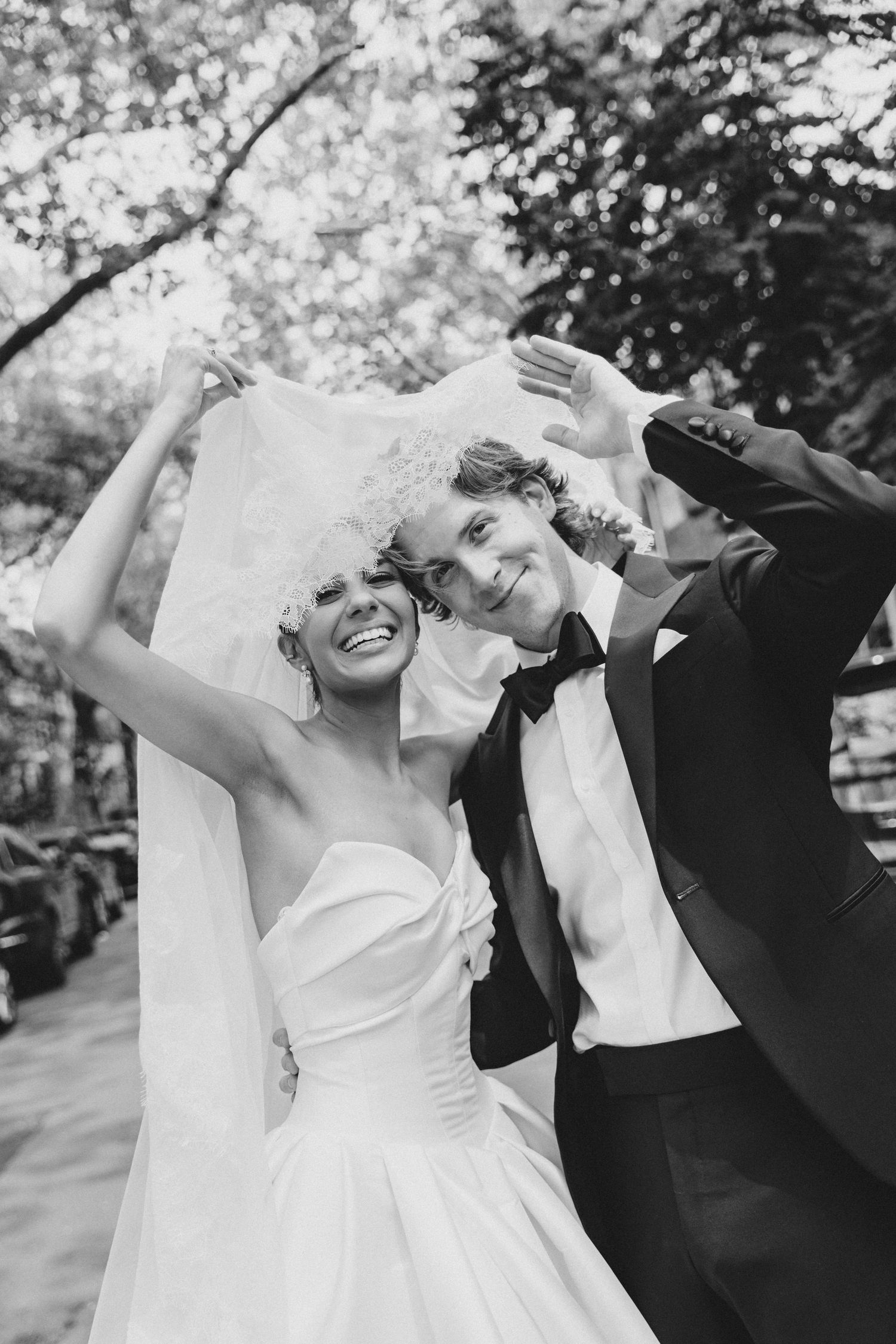 Black and white photo of joyful bride and groom celebrating outdoors, veil flowing, trees in background.