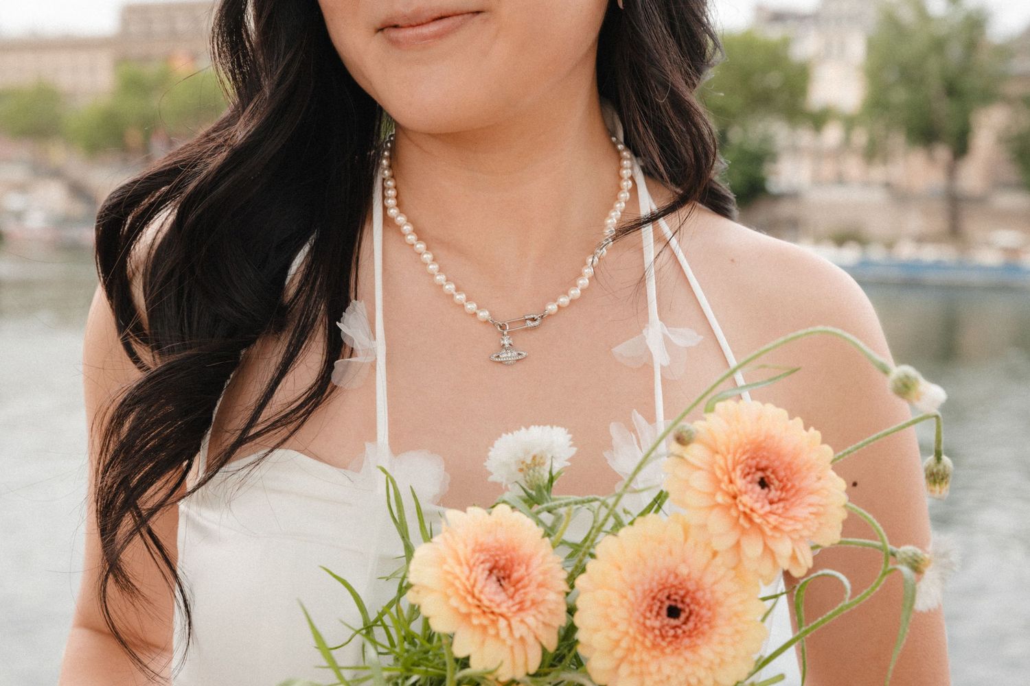 Bride holding peach dahlia bouquet wearing pearl necklace and white floral wedding dress near waterfront.