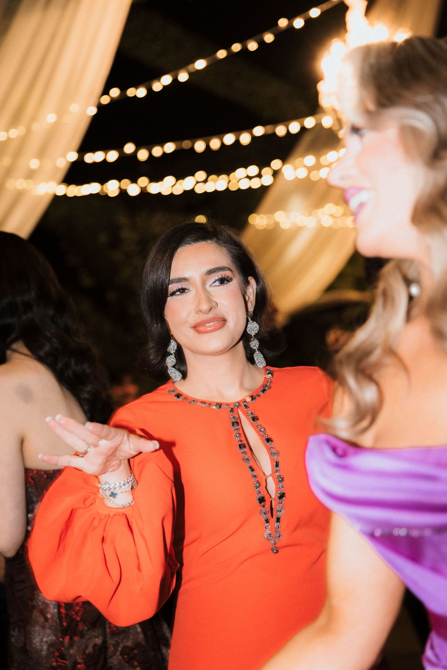 Woman in bright orange dress and sparkling jewelry at an evening event with twinkling lights in the background.