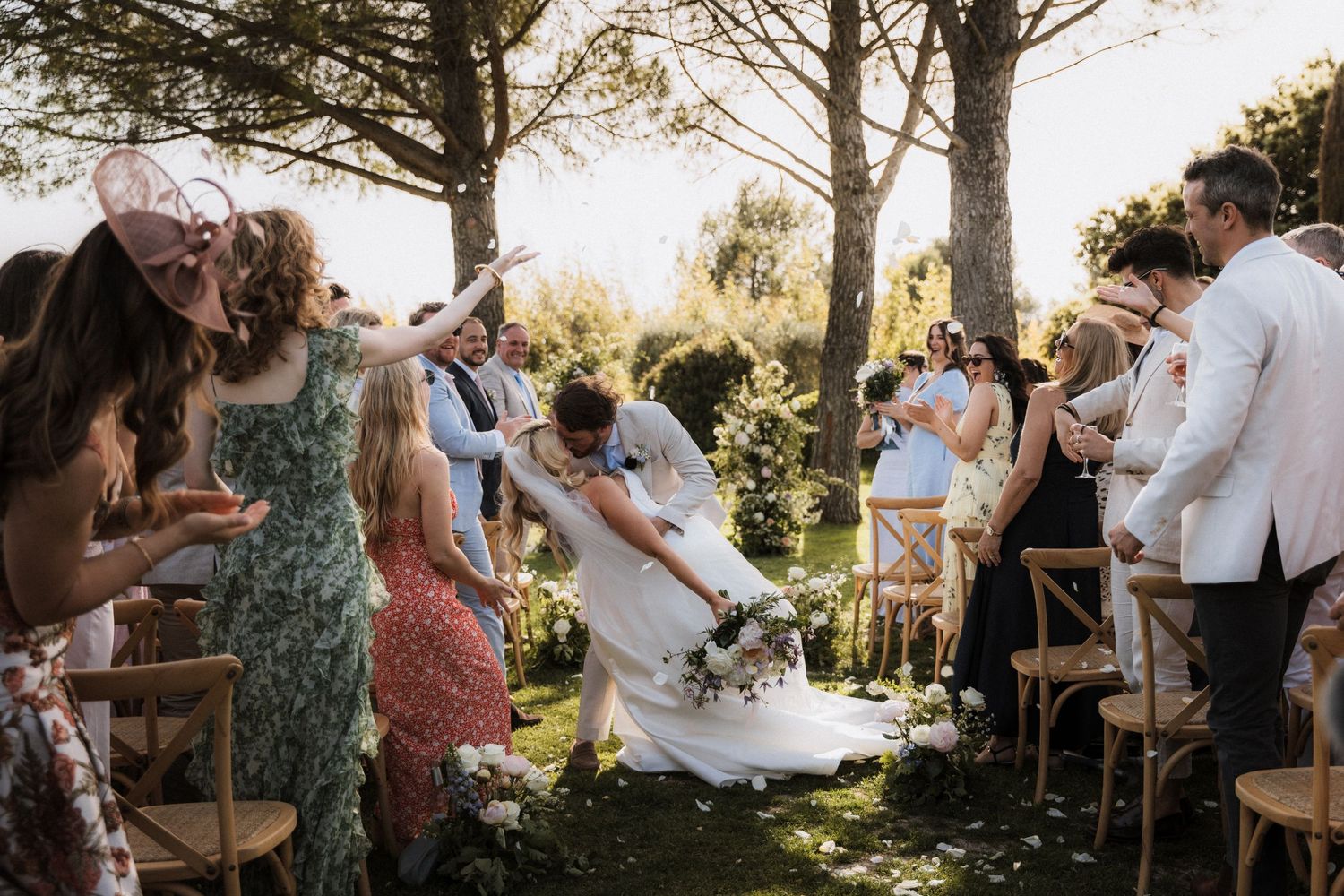 Wedding guests throw flower petals at a joyful outdoor ceremony under tall trees.
