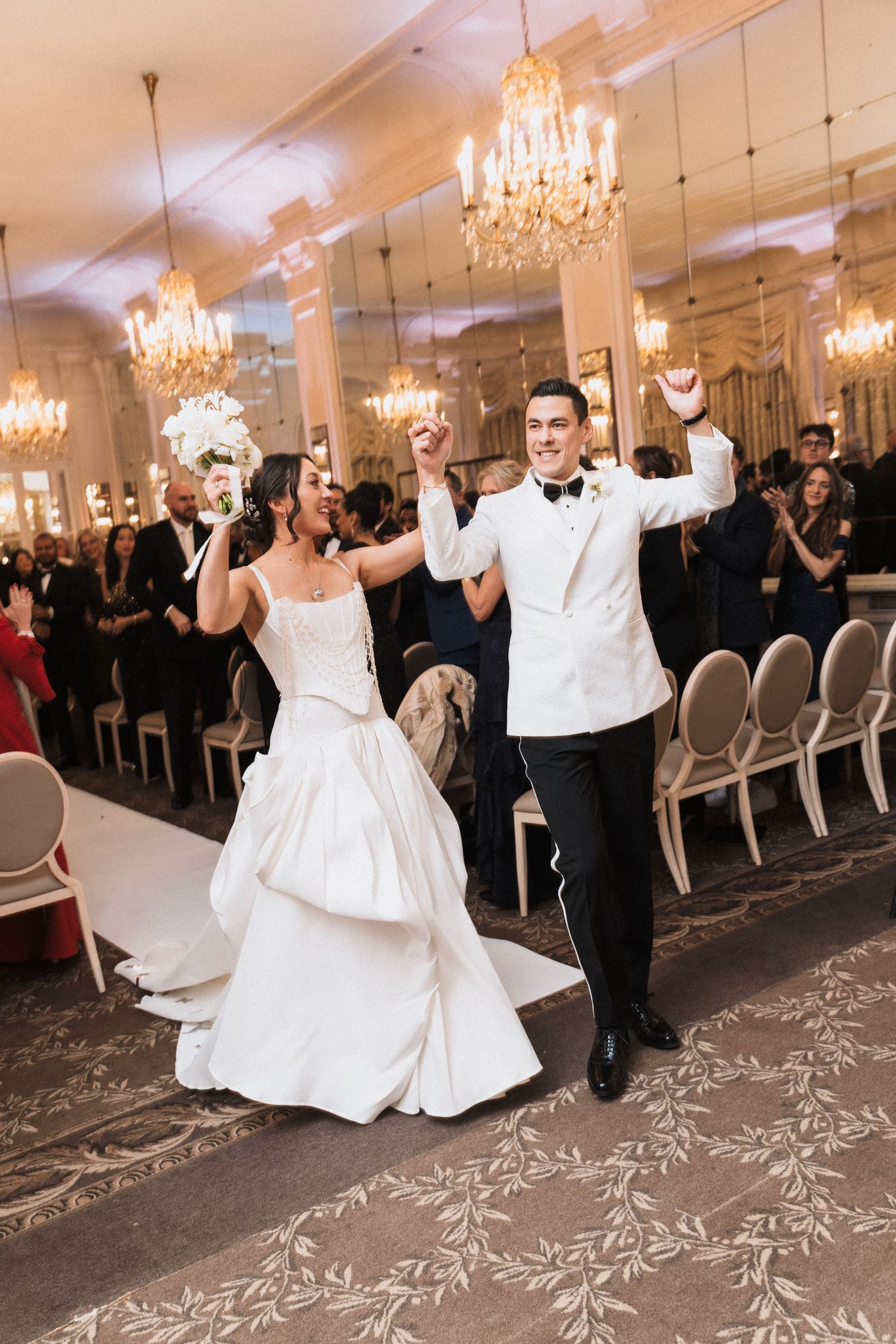 Bride and groom celebrate grand entrance at elegant ballroom wedding reception with chandeliers and guests applauding.