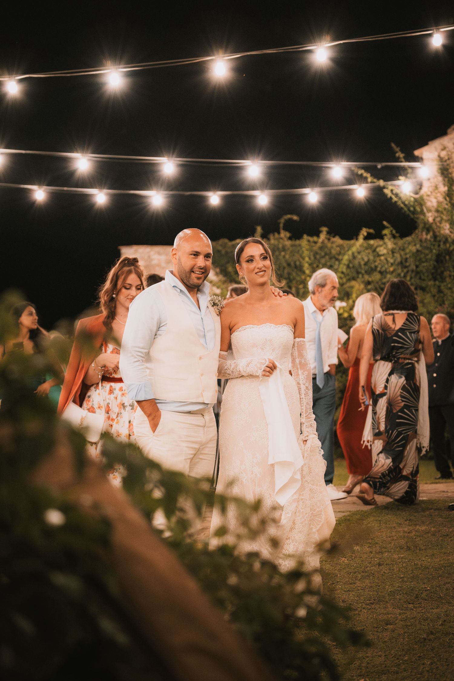 Bride and groom walking together at outdoor evening wedding reception under warm string lights.
