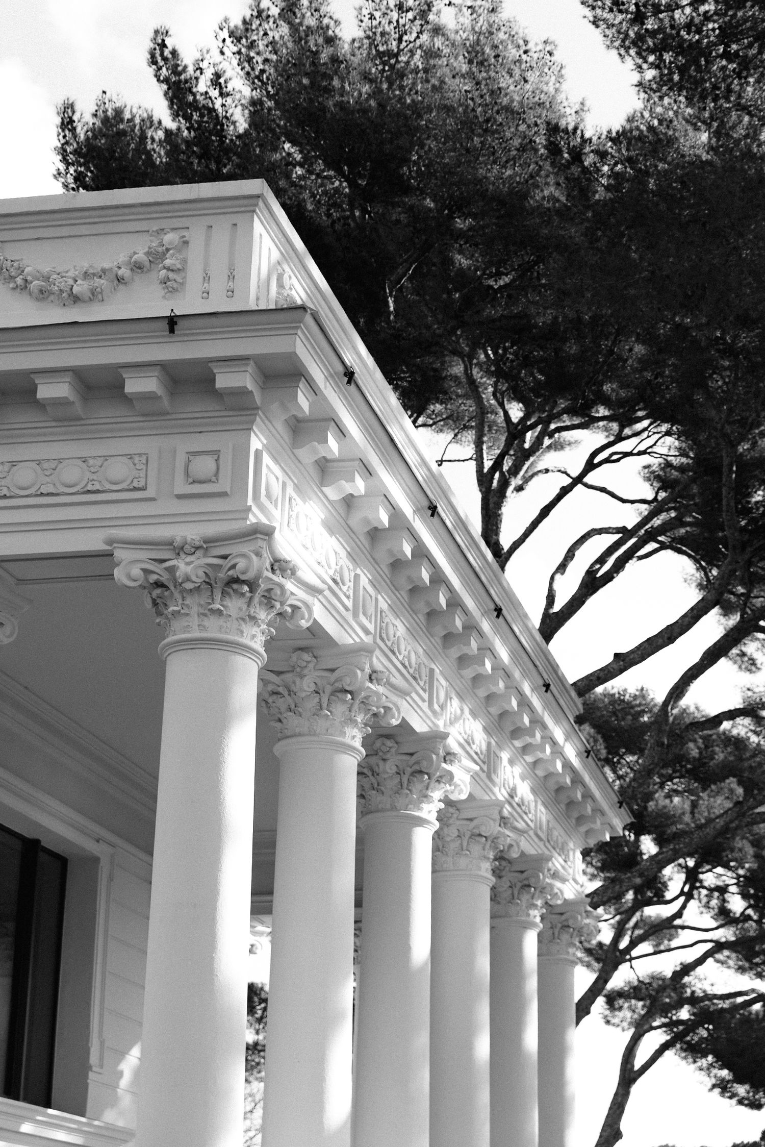 Villa Rocabella - Black and white photo of elegant neoclassical building with Corinthian columns, ornate frieze, & tall trees in background.