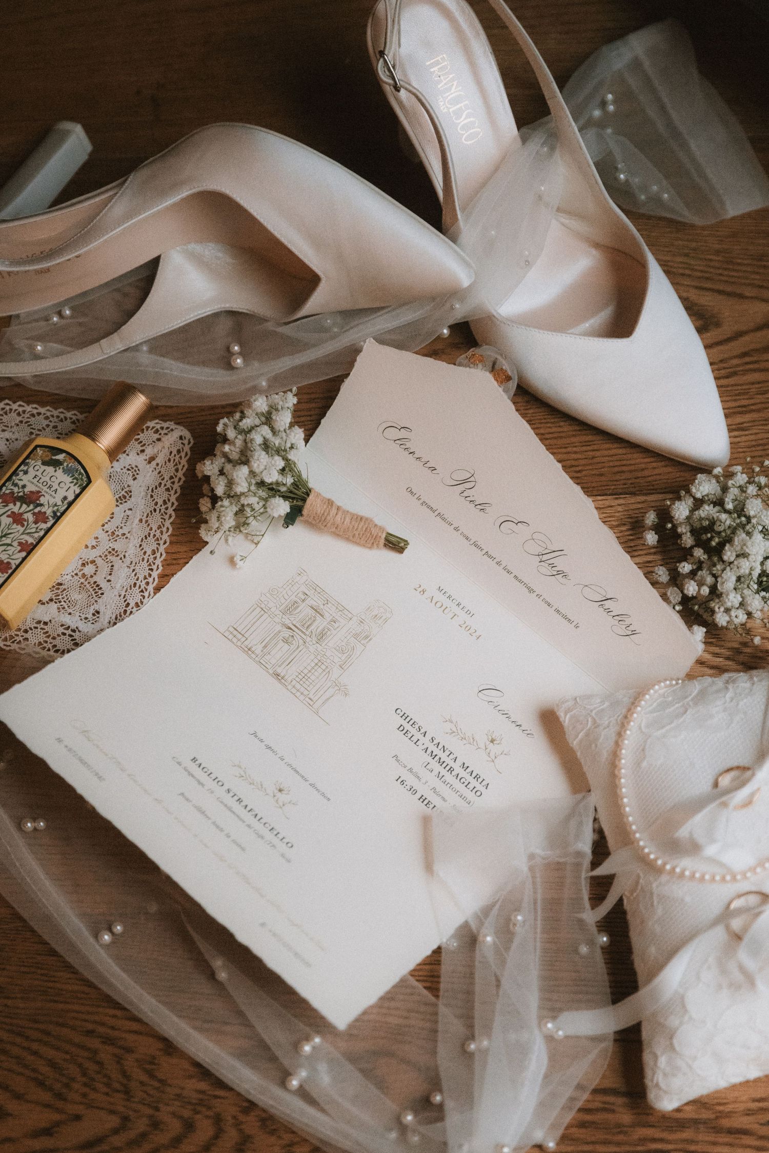 Elegant wedding flat lay featuring white heels, floral invitations, baby's breath, pearl accessories, and a sheer veil on wood.