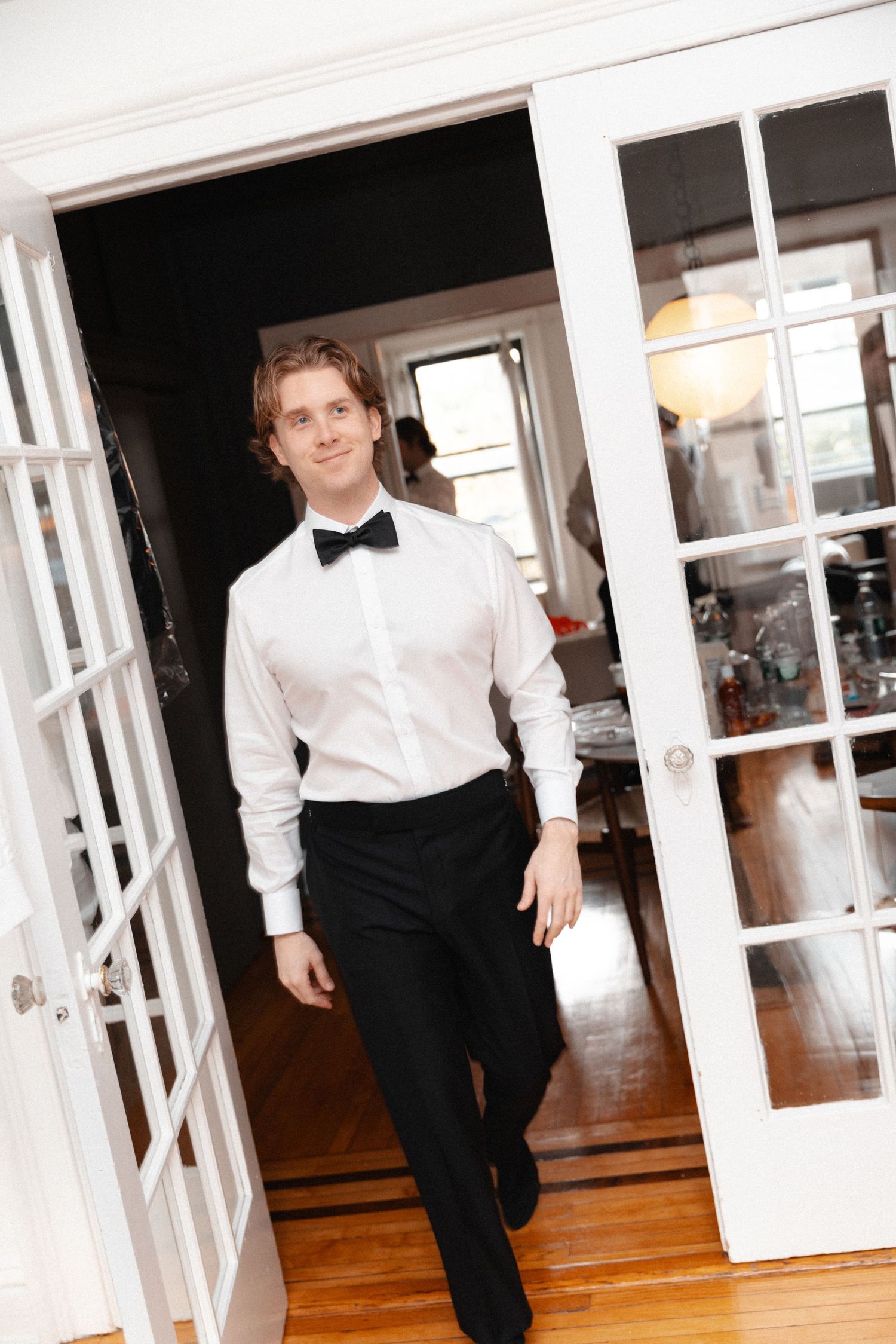 Young man in formal tuxedo shirt and bow tie walking through French doors in elegant home interior.