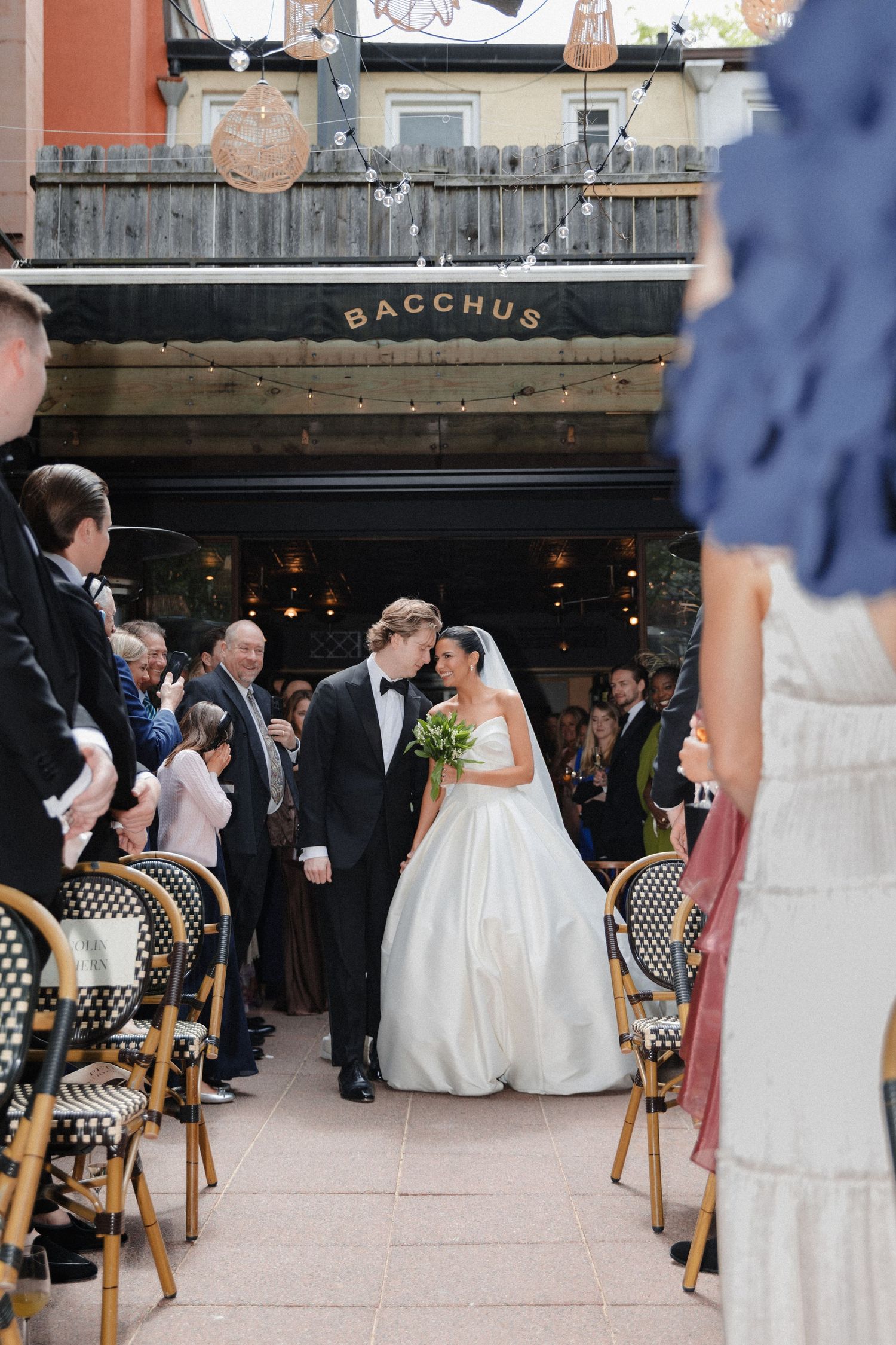 Bride and groom share a kiss walking down the aisle outside Bacchus restaurant, surrounded by cheering wedding guests.