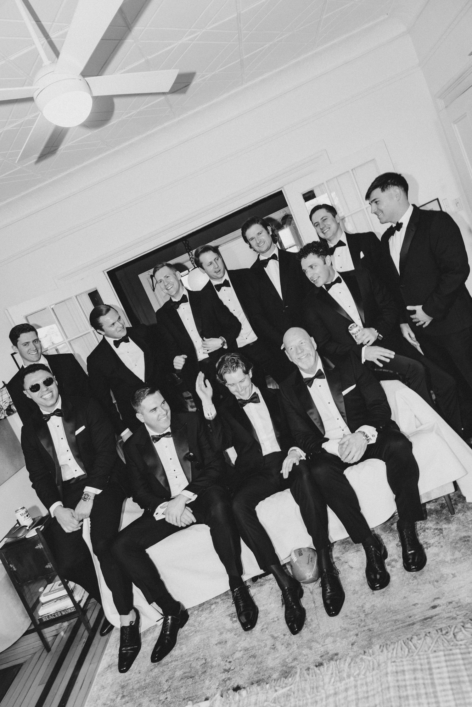 Black and white photo of groomsmen in tuxedos posing together, some seated and some standing, smiling at a wedding.