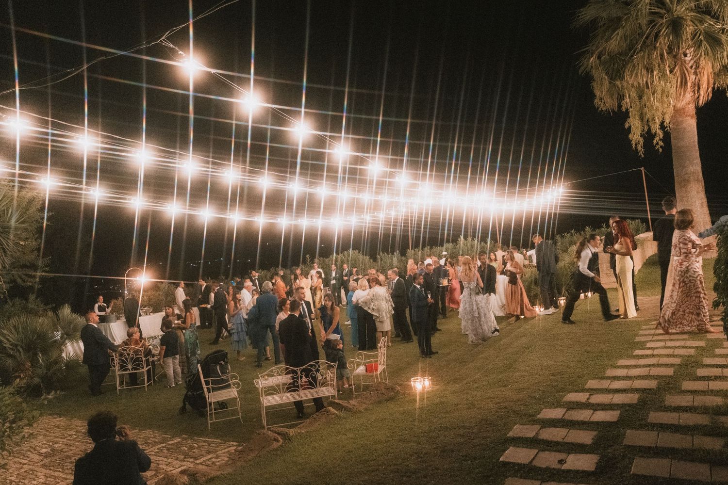 Outdoor evening wedding reception with string lights overhead, guests mingling on grass under warm glowing bistro lights.