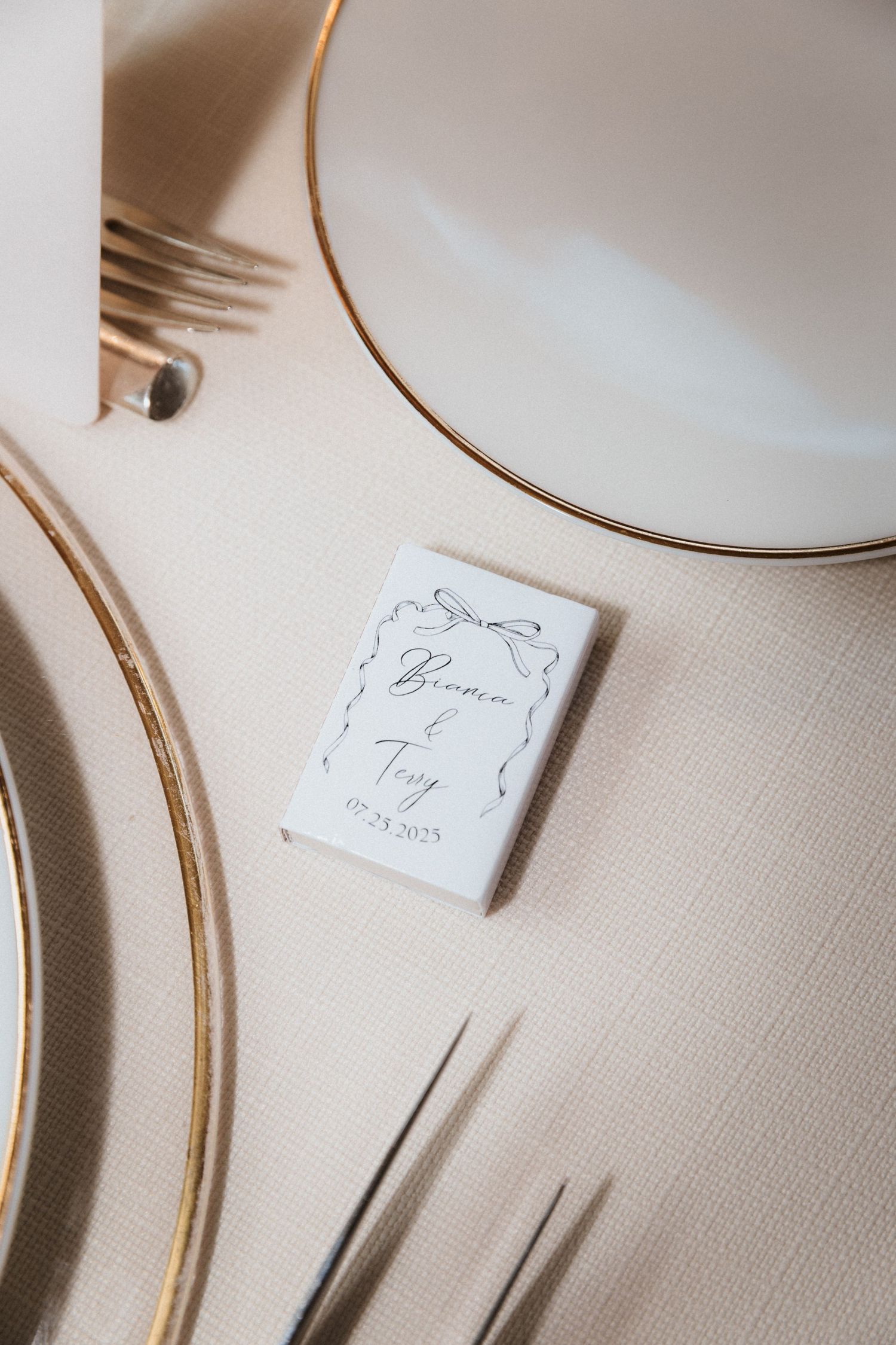 Wedding place card with elegant script text sits on a cream tablecloth beside gold-rimmed plates.