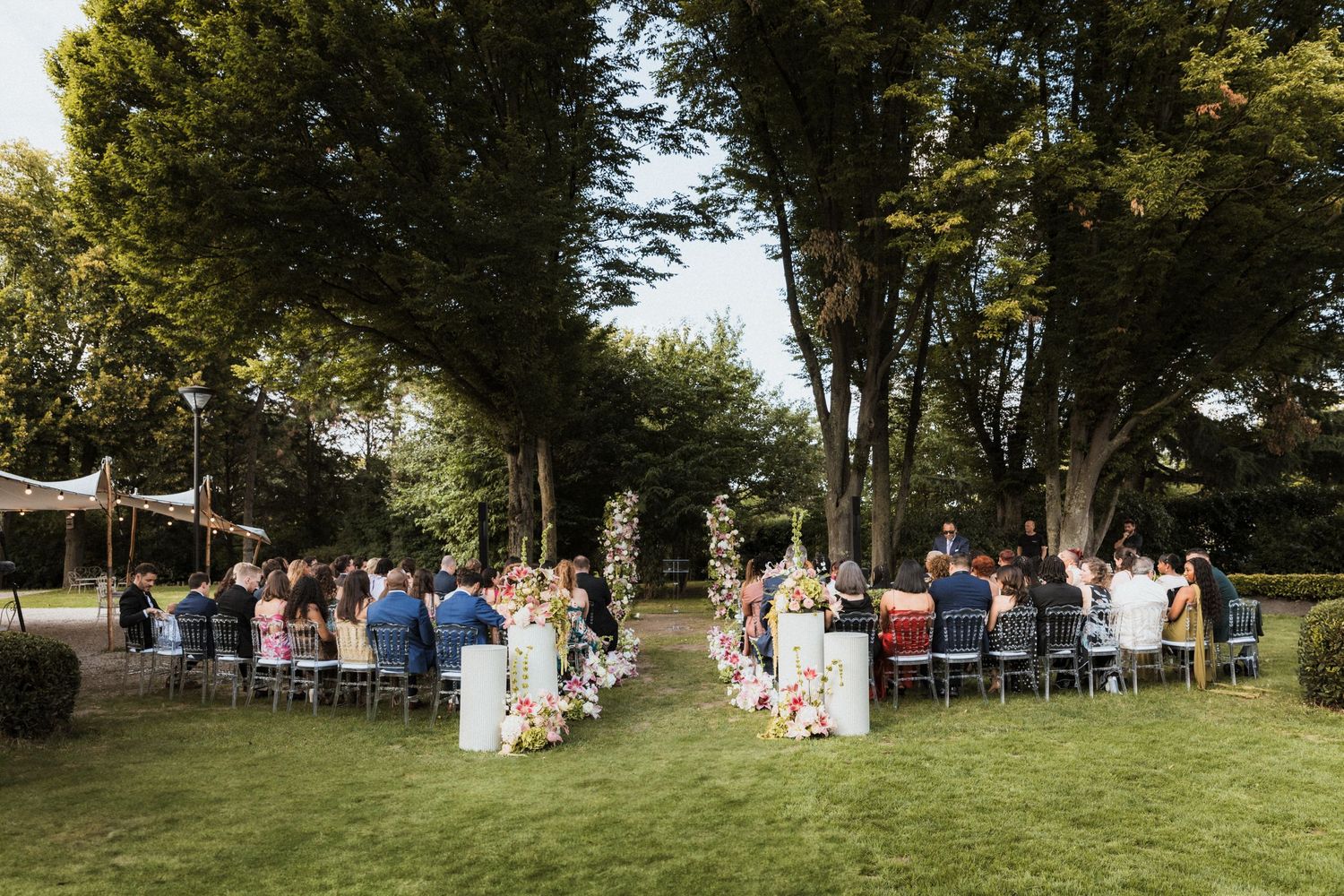 Garden wedding ceremony setup with white chairs and floral arch under tall trees.
