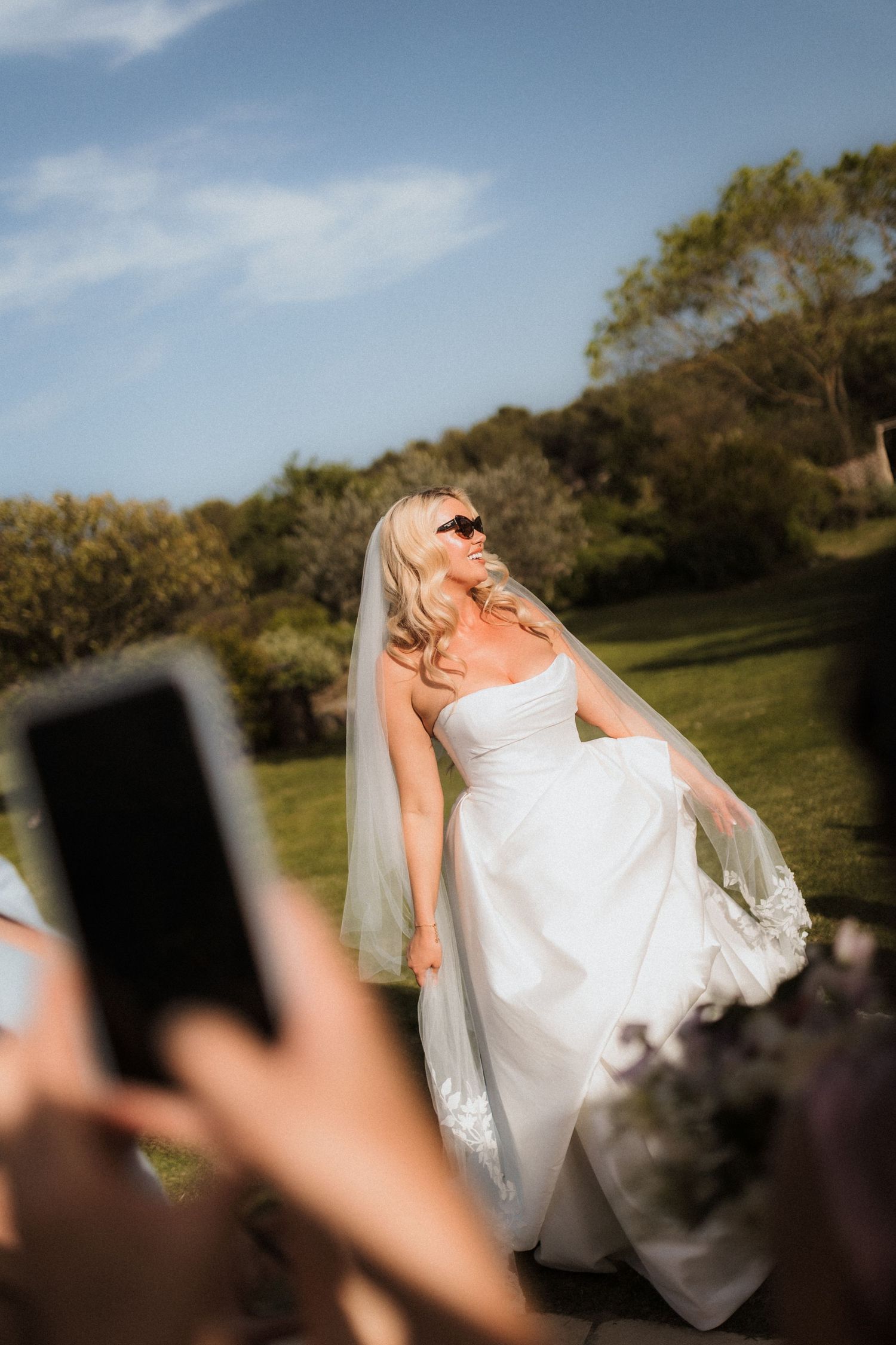 Bride in flowing white dress twirling outdoors against natural landscape and blue sky.