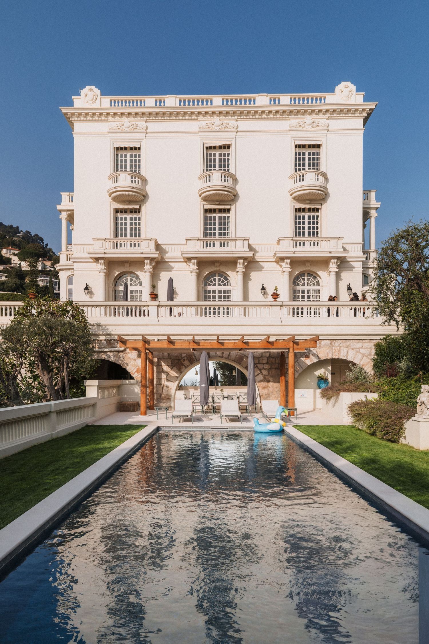 Elegant neoclassical Villa La Vigie, Monaco with a rectangular reflecting pool, manicured lawn, and wooden pergola under a clear blue sky.
