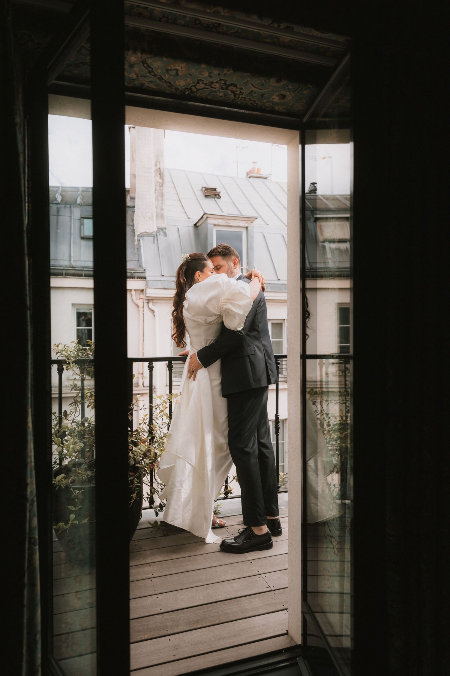 A romantic couple shares an intimate moment by a window in a dimly lit room with urban architecture visible outside.