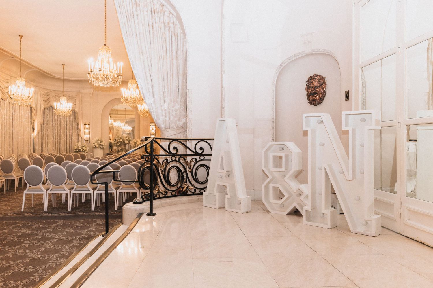 Elegant white ballroom interior with crystal chandeliers and ornate wrought iron railings along a curved staircase.