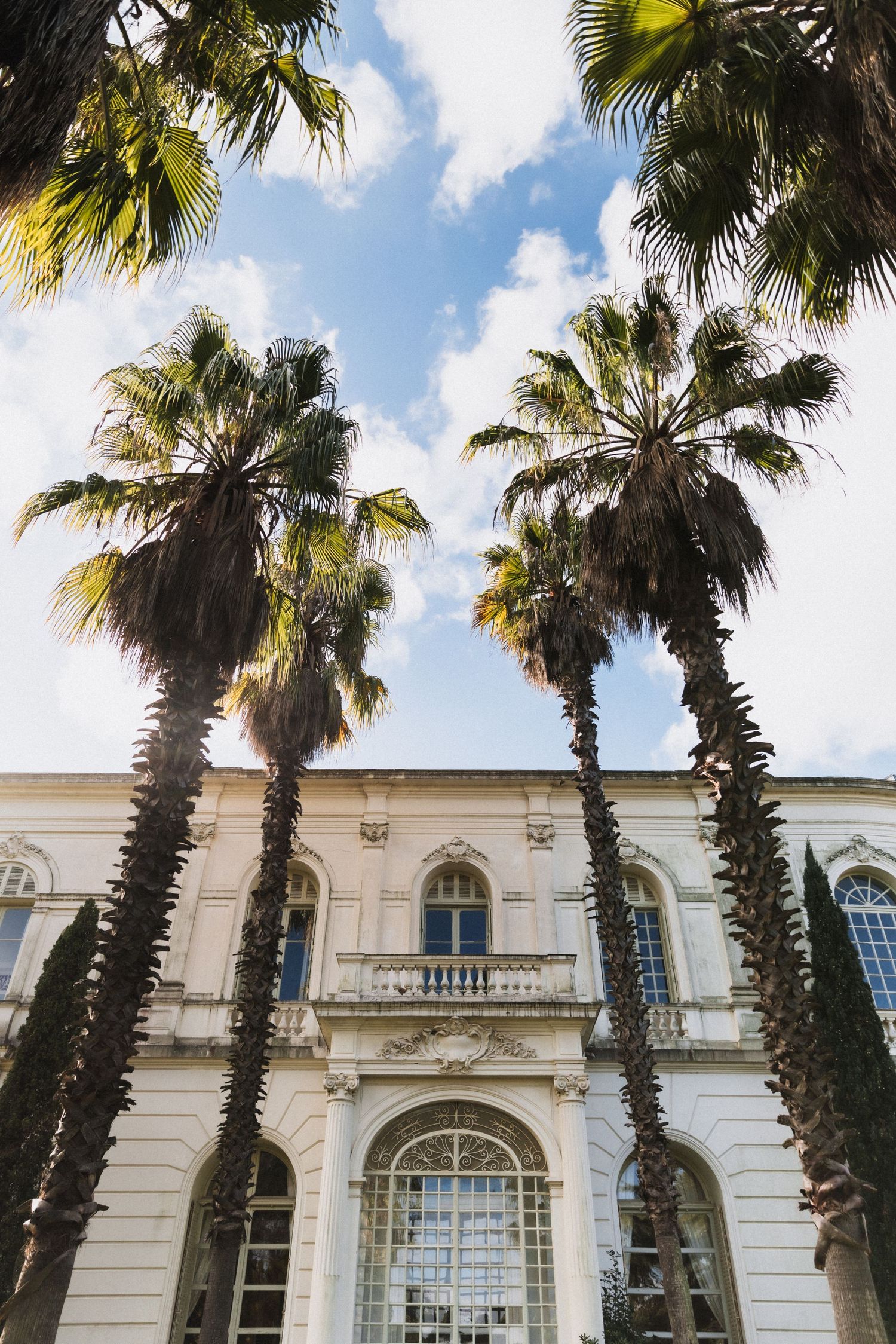 Tall palm trees frame a grand white neoclassical building facade with arched windows and balcony under a partly cloudy sky.