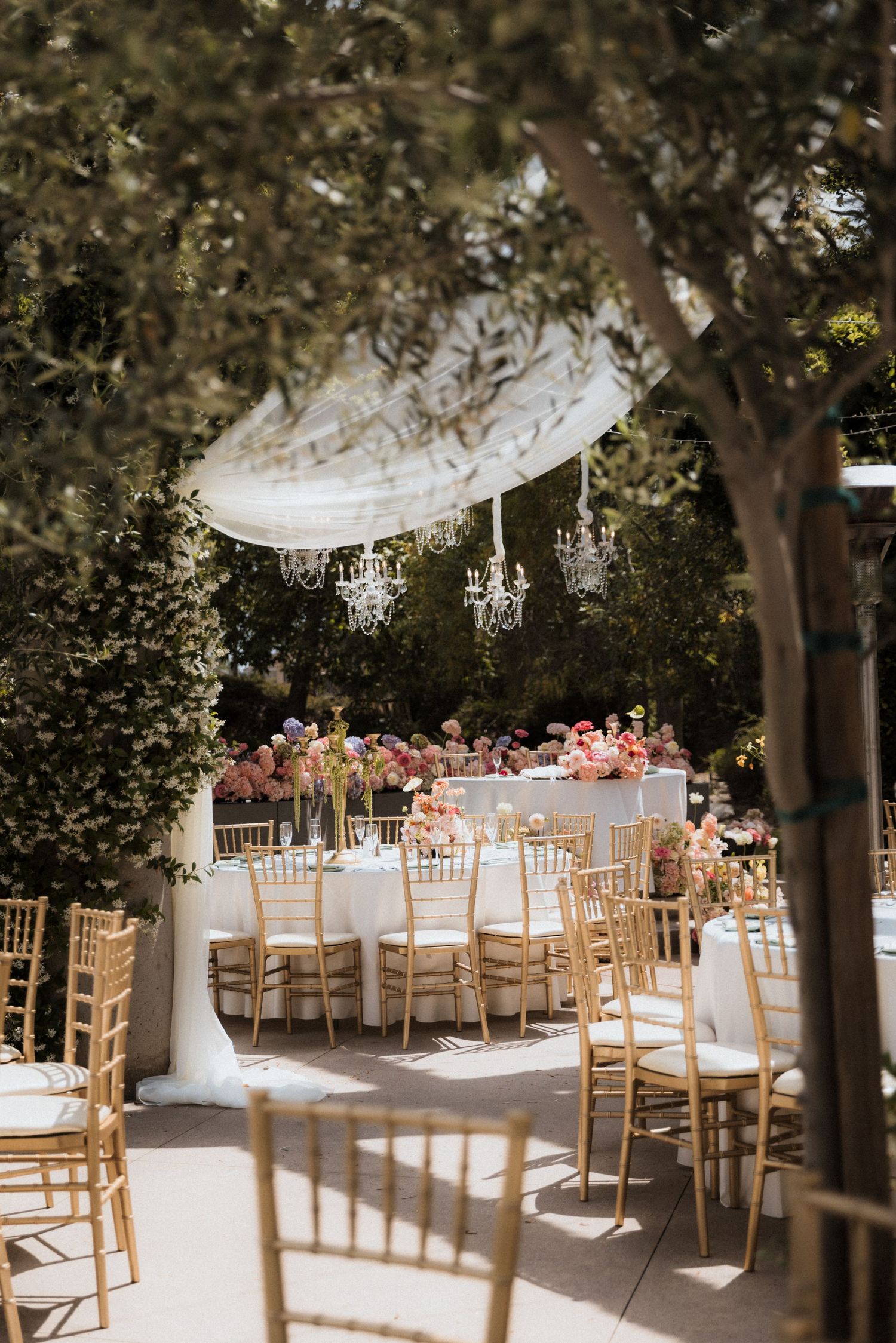 Elegant outdoor wedding reception with gold chiavari chairs, crystal chandeliers, and floral centerpieces under a draped canopy.