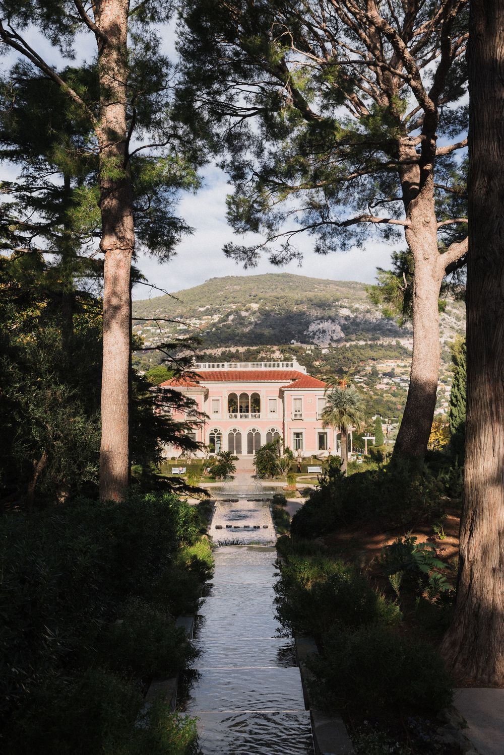 Elegant pink villa framed by tall pine trees with a reflective garden canal and mountain backdrop in southern France.