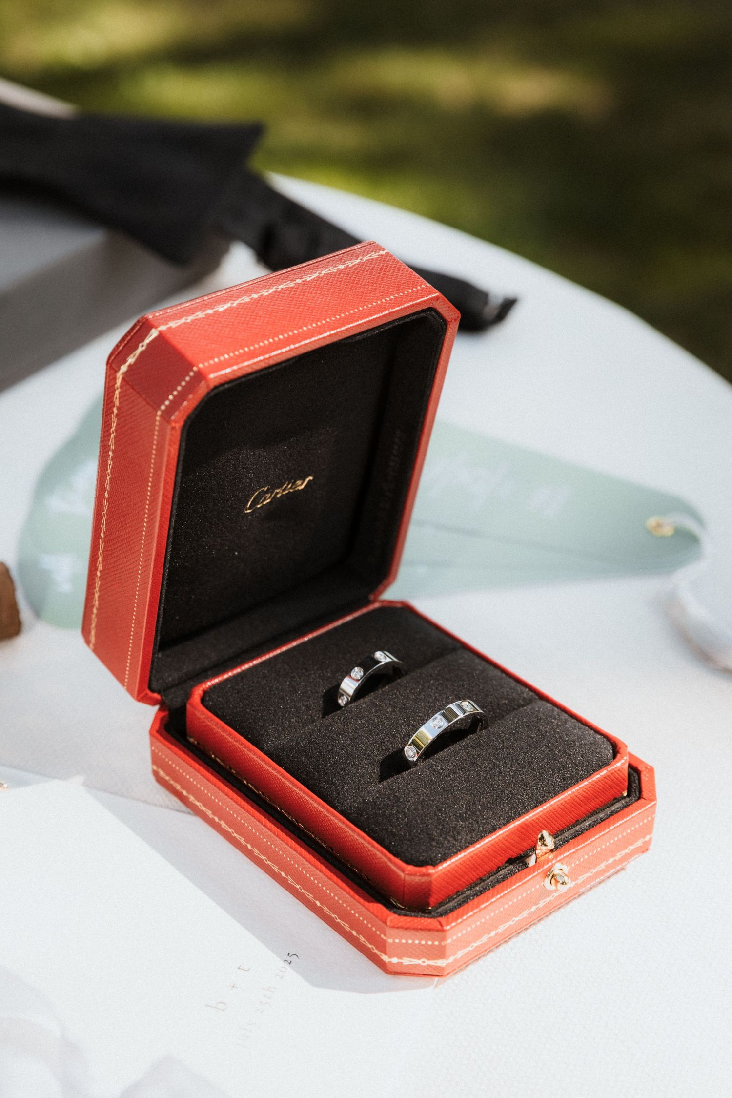 Two silver wedding bands displayed in an open red Cartier jewelry box on a white table outdoors.
