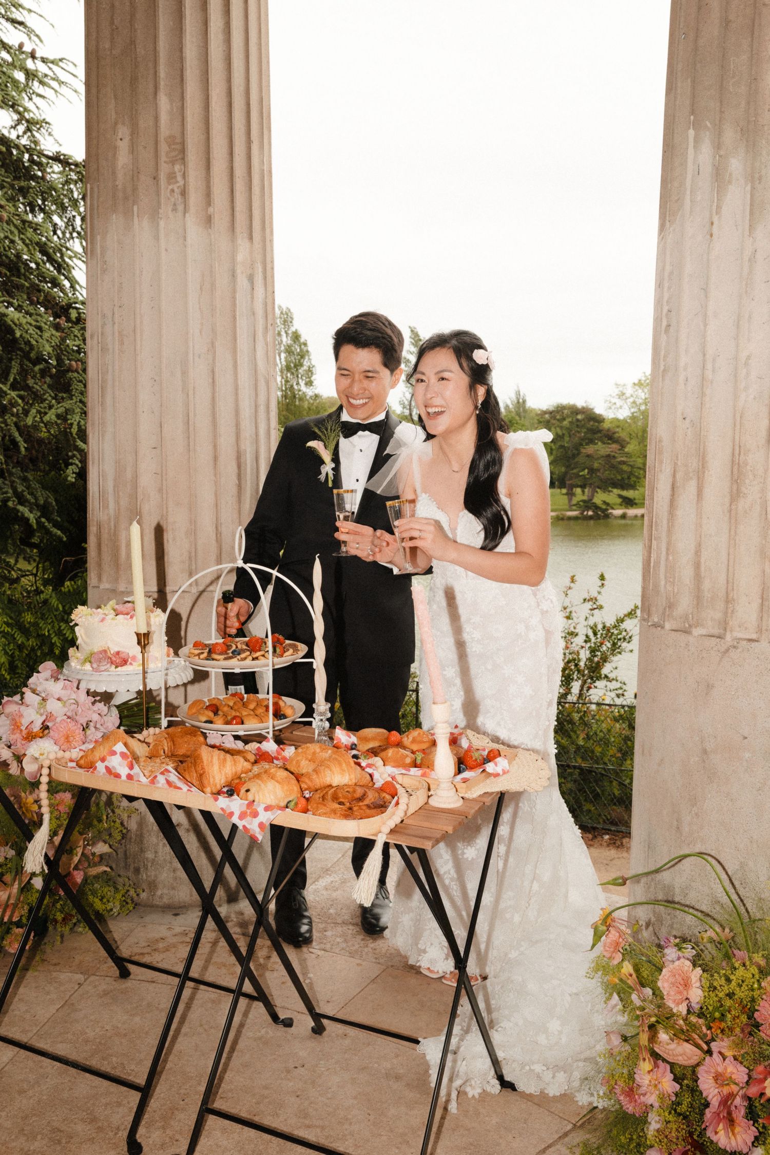 A bride and groom toast with champagne beside an elegant outdoor dessert table with croissants and flowers near a lake.