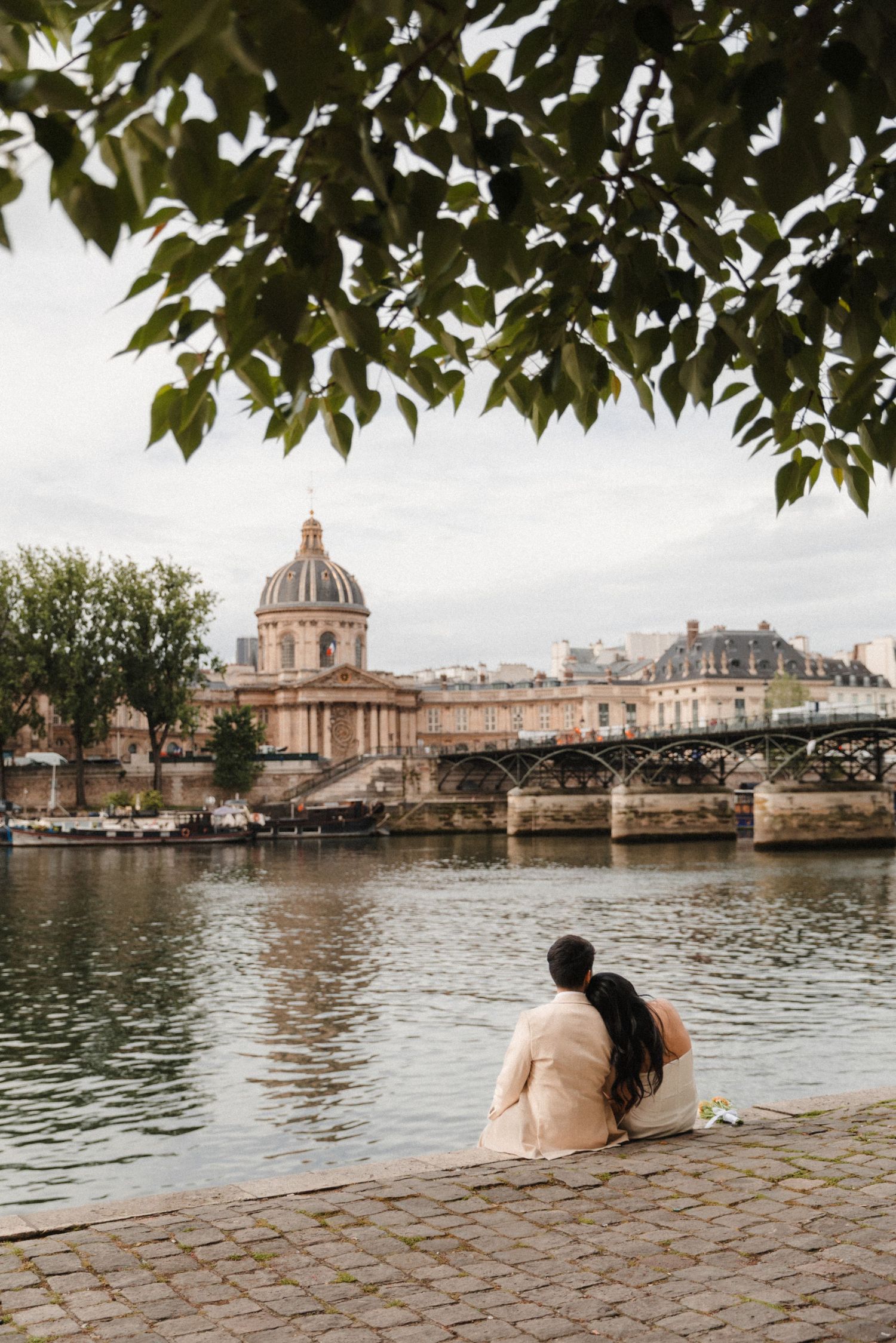 Couple sitting by the Seine River in Paris, with Pont des Arts bridge and Institut de France dome in background.