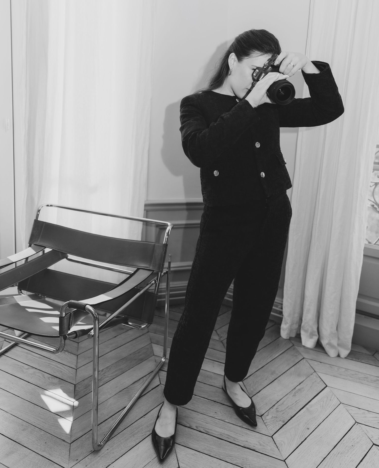 Black and white photo of a woman in a black suit photographing with a camera in a minimalist room with herringbone floors.