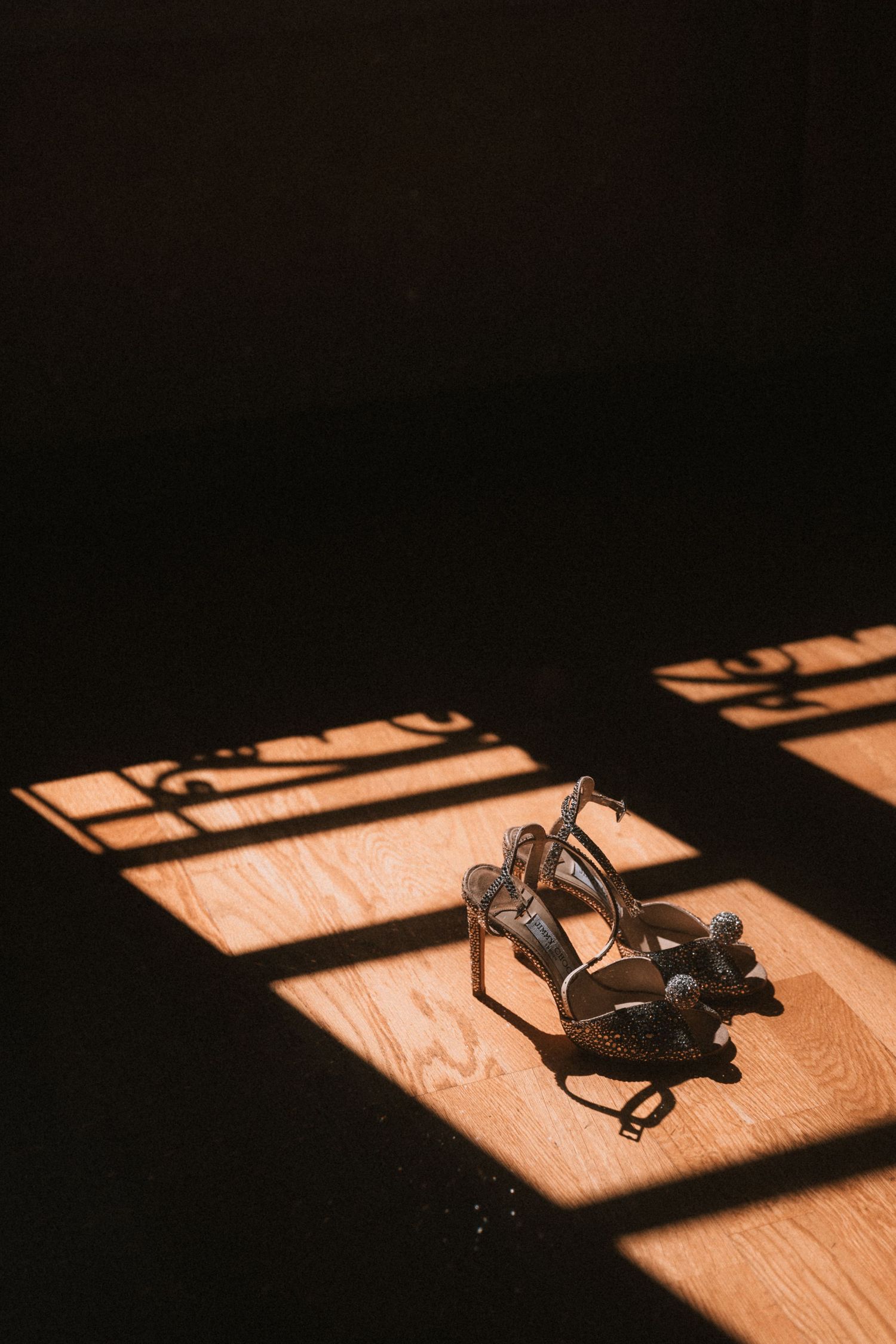Dramatic lighting on wedding shoes casting shadows on wooden floor.