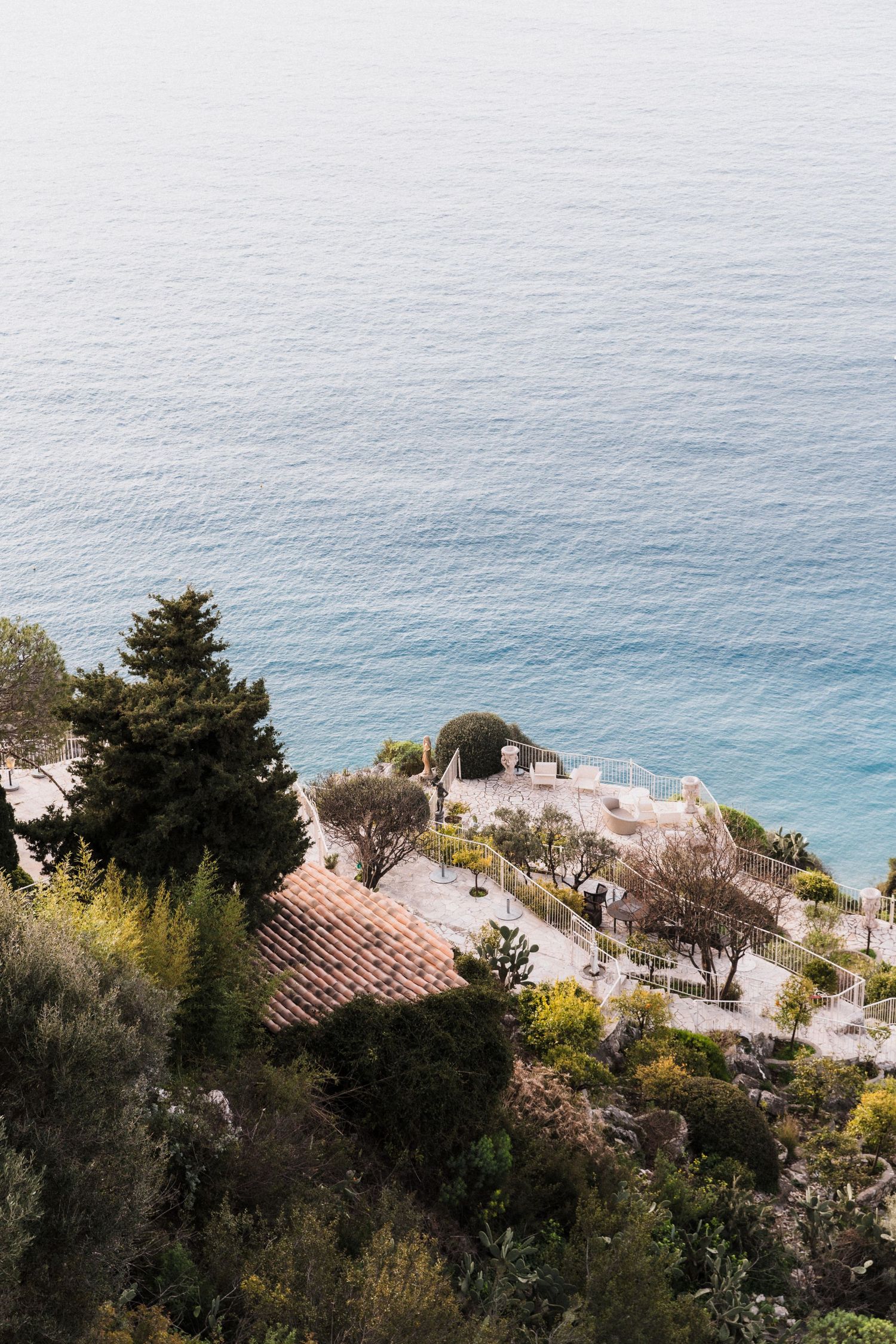 Château Eza - Aerial view of a Mediterranean coastal garden with terracotta rooftops overlooking a calm blue sea.