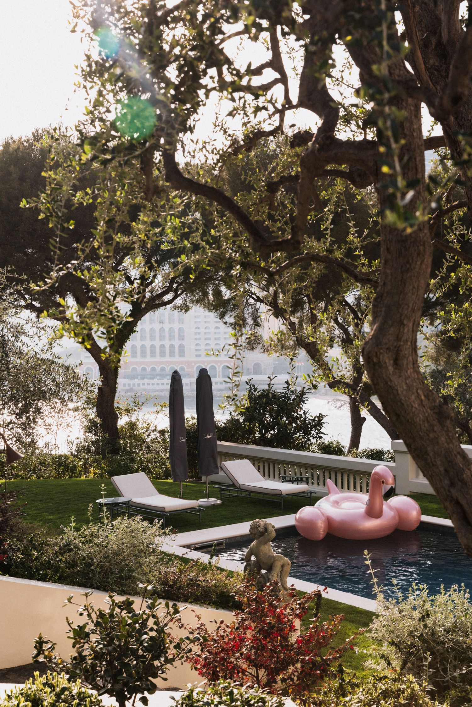 Villa La Vigie, Monaco - Luxury poolside garden with pink flamingo float, lounge chairs, and large trees overlooking a waterfront cityscape.