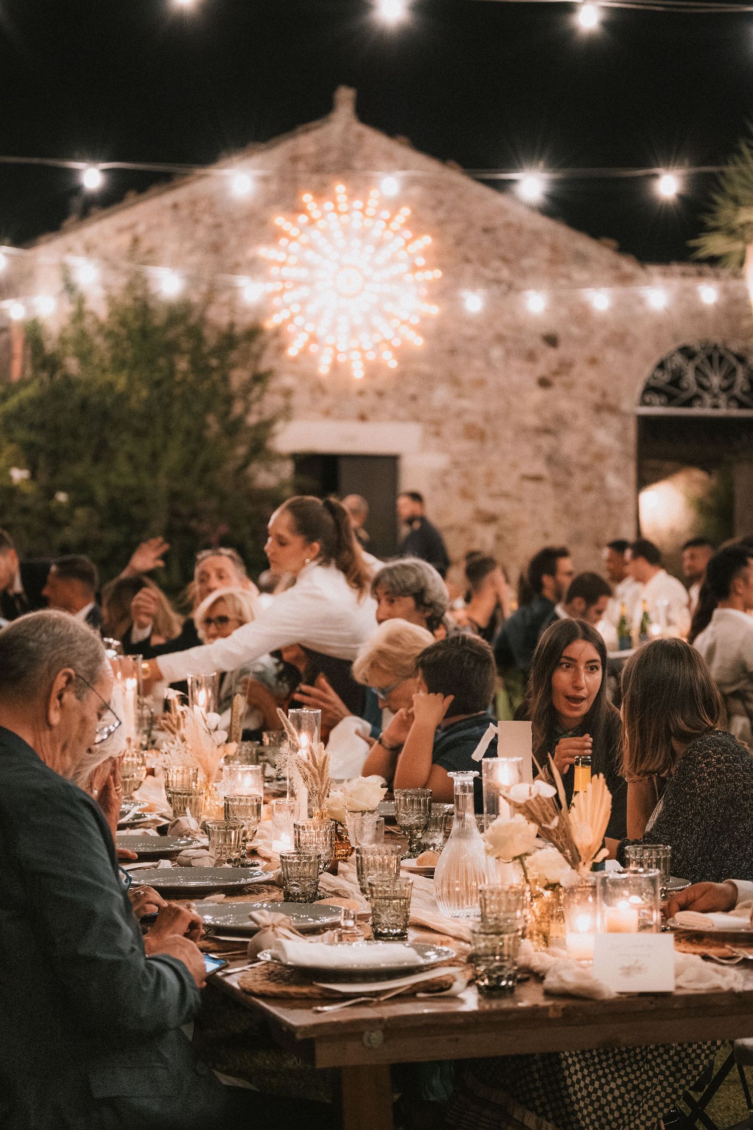 Outdoor wedding reception at dusk with stone building backdrop, string lights, and candlelit tables.