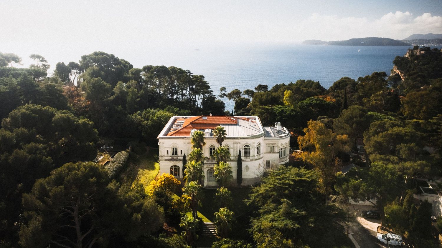 Luxurious white villa nestled among trees overlooking a Mediterranean coastline with blue waters and mountains.