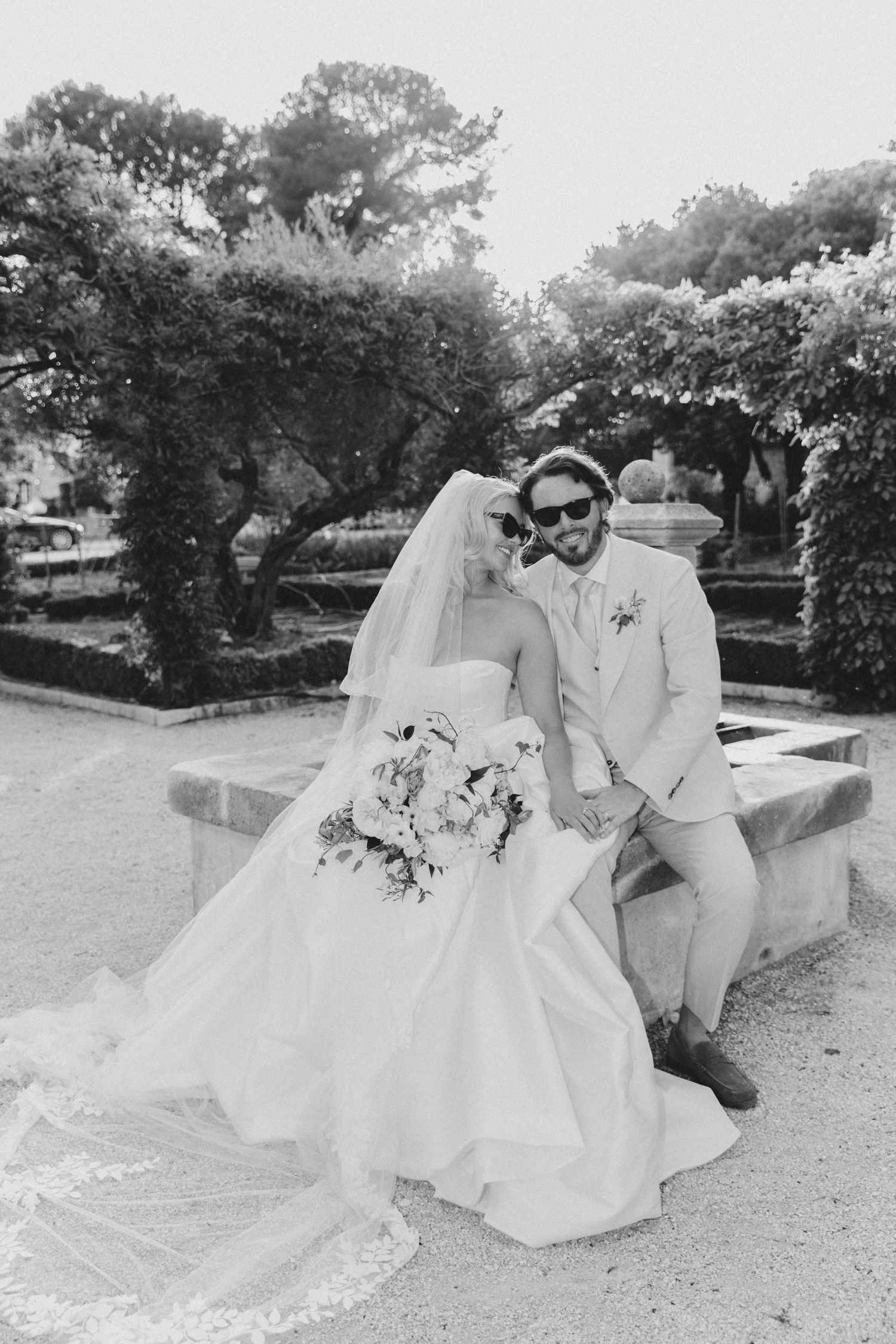 A romantic wedding photoshoot featuring a couple sitting together on a stone bench in a garden setting.