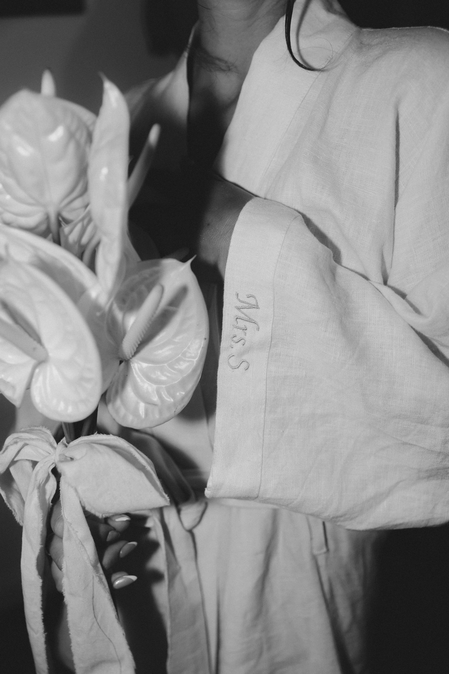 Black and white photo of a person in a linen robe holding white anthurium flowers with embroidered monogram detail.