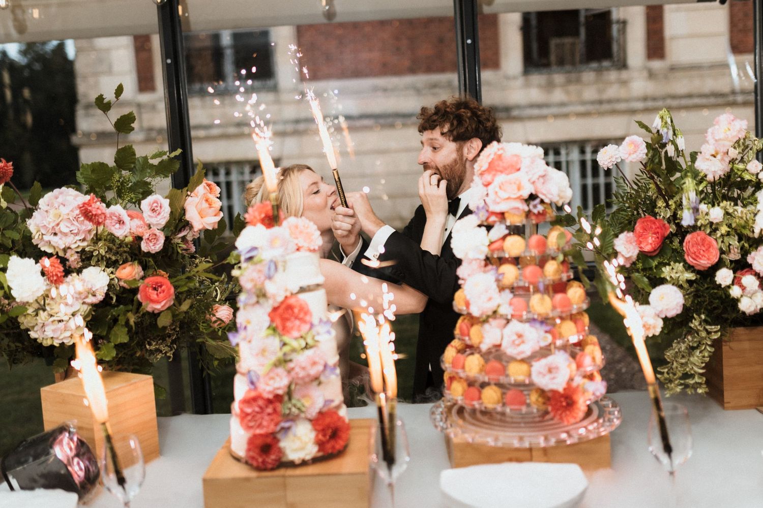 Bride and groom cutting a floral wedding cake with sparklers and a croquembouche on an elegant decorated table.