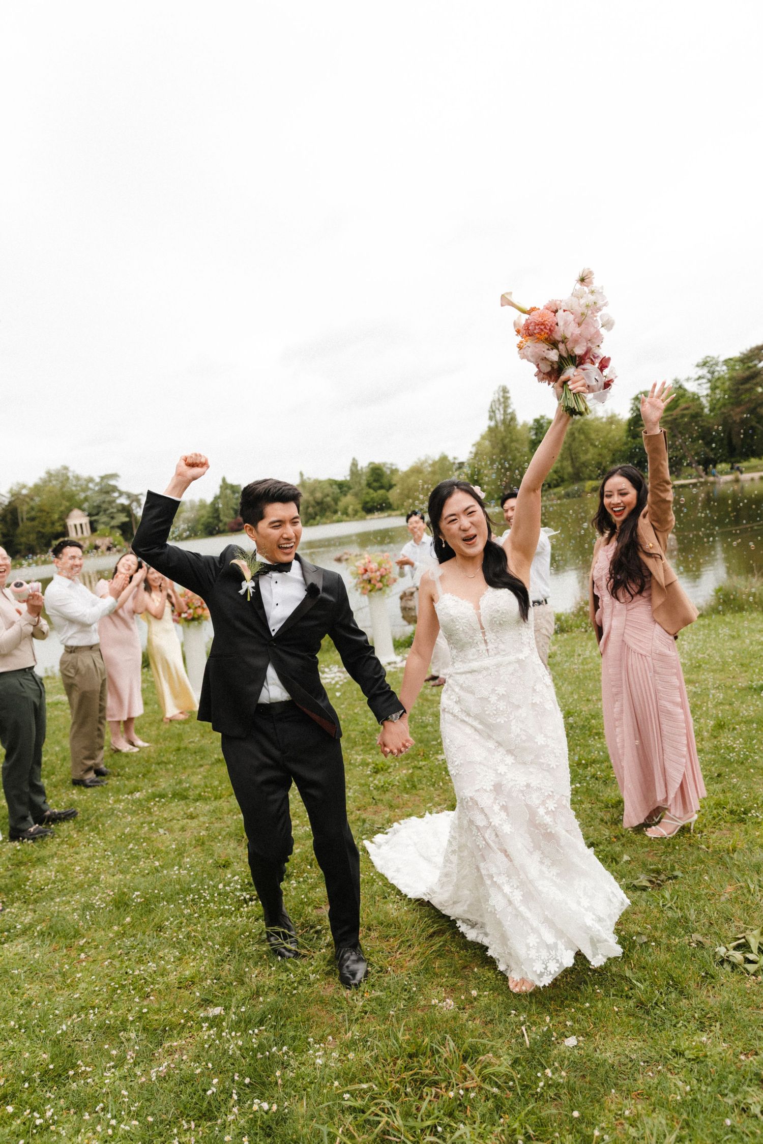 Joyful newlywed Asian couple celebrating after wedding ceremony outdoors, bride in lace gown holding bouquet, groom in black tuxedo.
